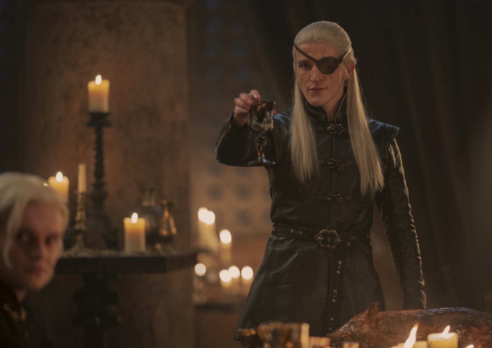 Actor Ewan Mitchell who plays an older Aemond Targaryen in 'House of the Dragon.' He's wearing an eye patch, has long, blonde hair, and his holding up a wine glass.