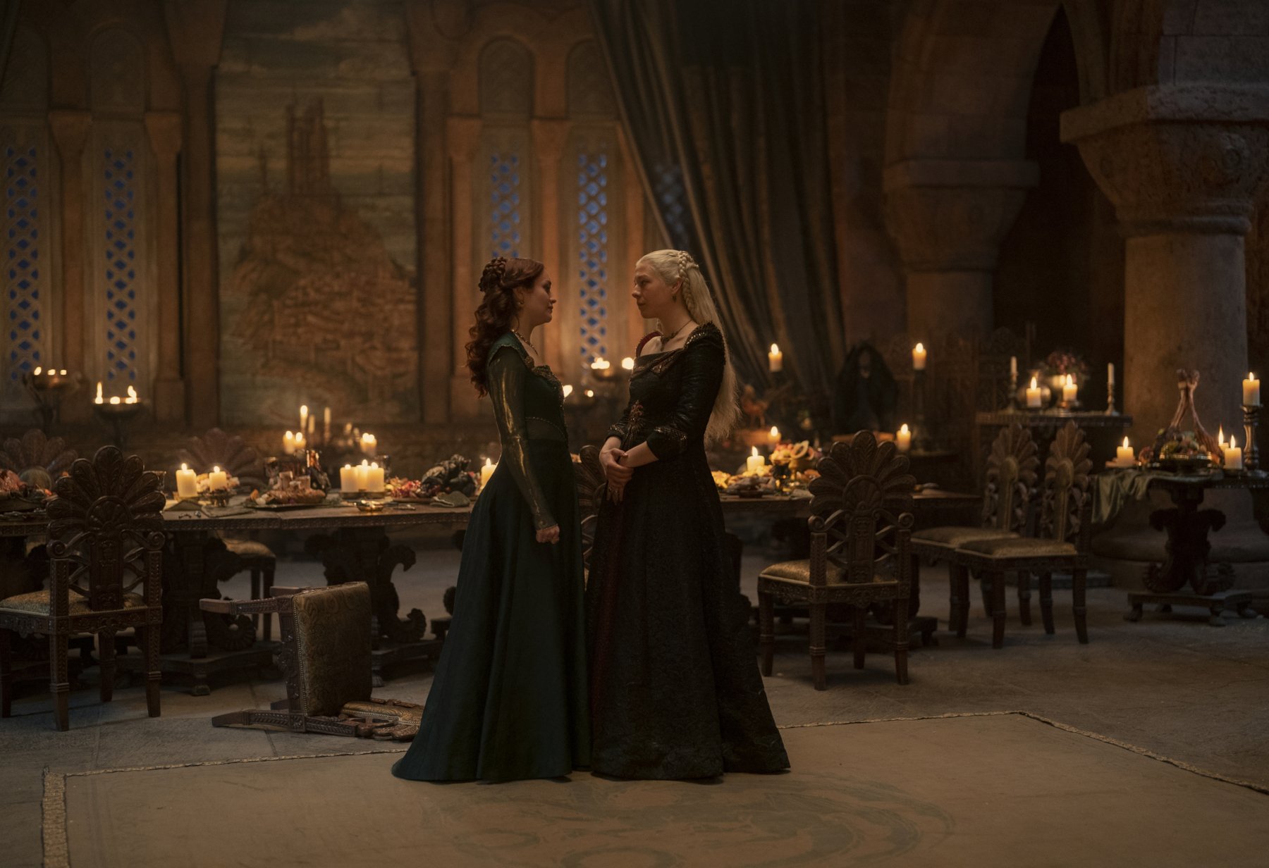Olivia Cooke and Emma D'Arcy as Alicent Hightower and Rhaenyra Targaryen in 'House of the Dragon' for our article about episode 9's release date on HBO. The two are facing one another in front of a dinner table, and they're holding onto each other.