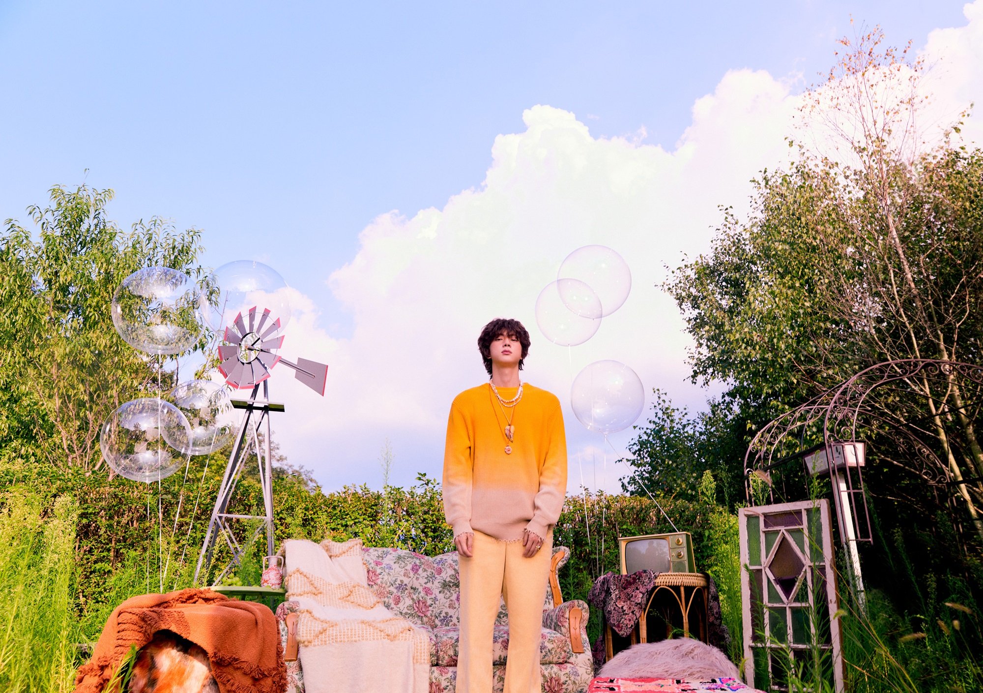 Jin of BTS stands outside in front of a floral couch and vintage TV