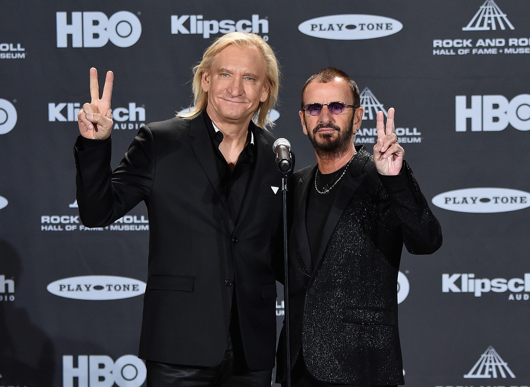 Joe Walsh and Ringo Starr stand in front of a microphone and hold up peace signs.