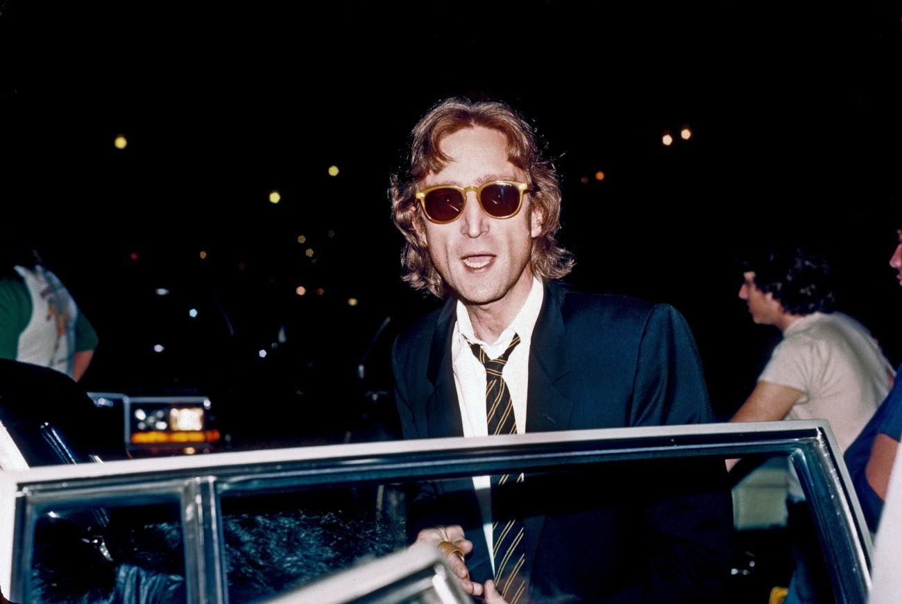 John Lennon wears sunglasses and stands in front of an open car door.