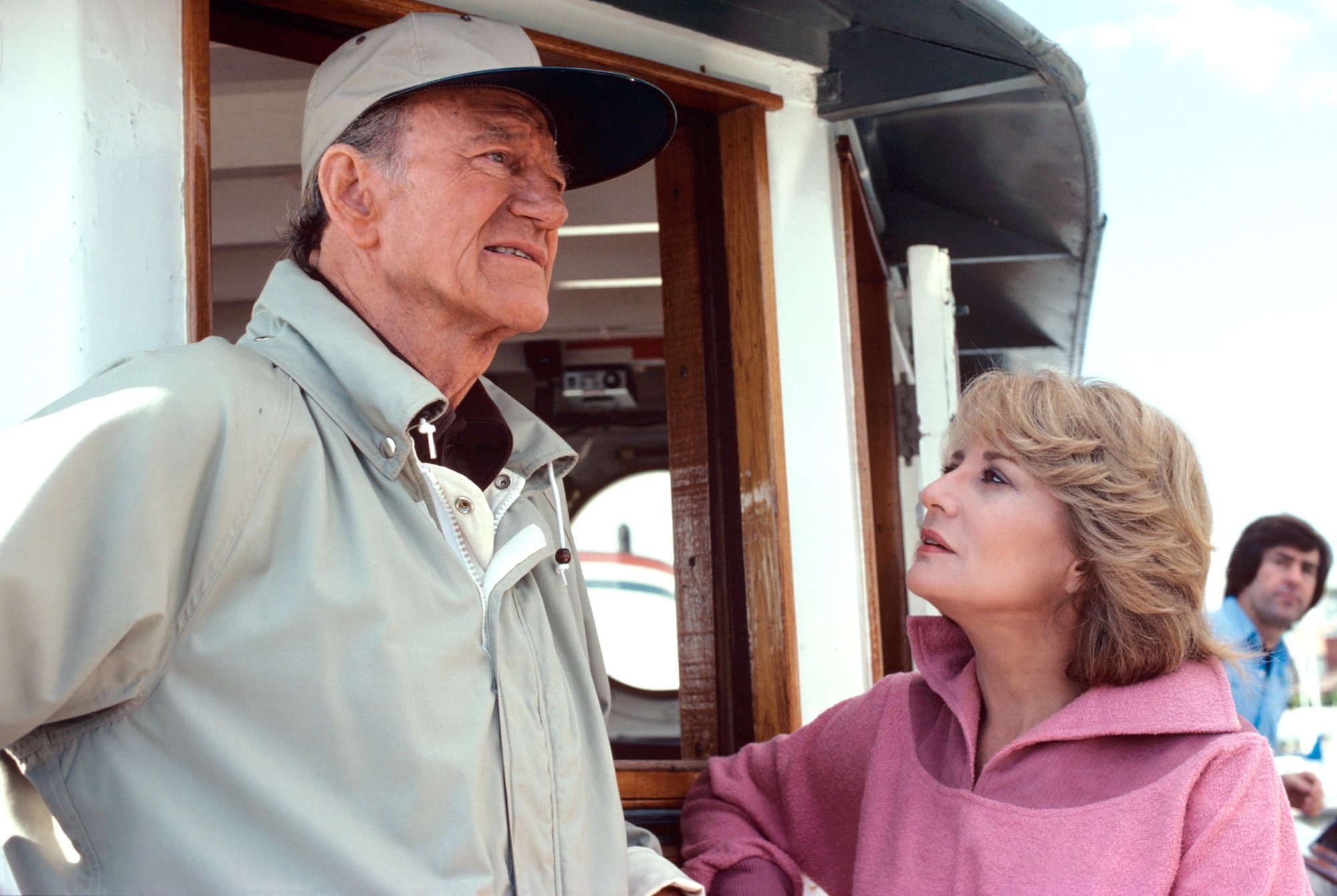 John Wayne and Barbara Walters. They're wearing jackets on his boat. Walters is looking up at Wayne, who is looking out at the water.