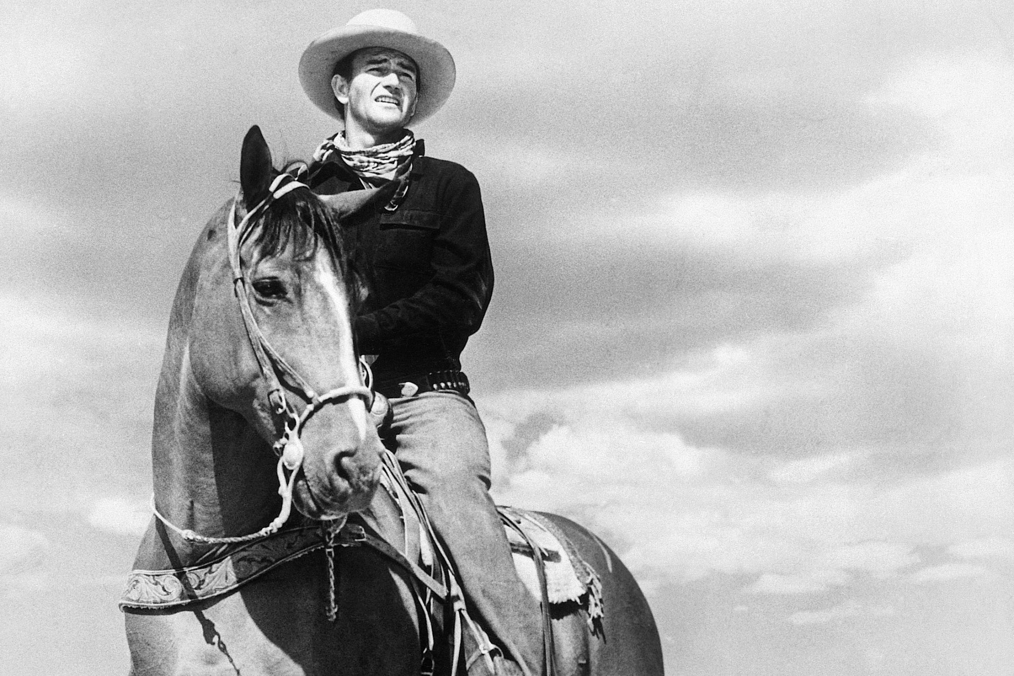 John Wayne starring in one of his Western movies. He's wearing a Western costume and cowboy hat in a black-and-white photo. He's riding a horse.