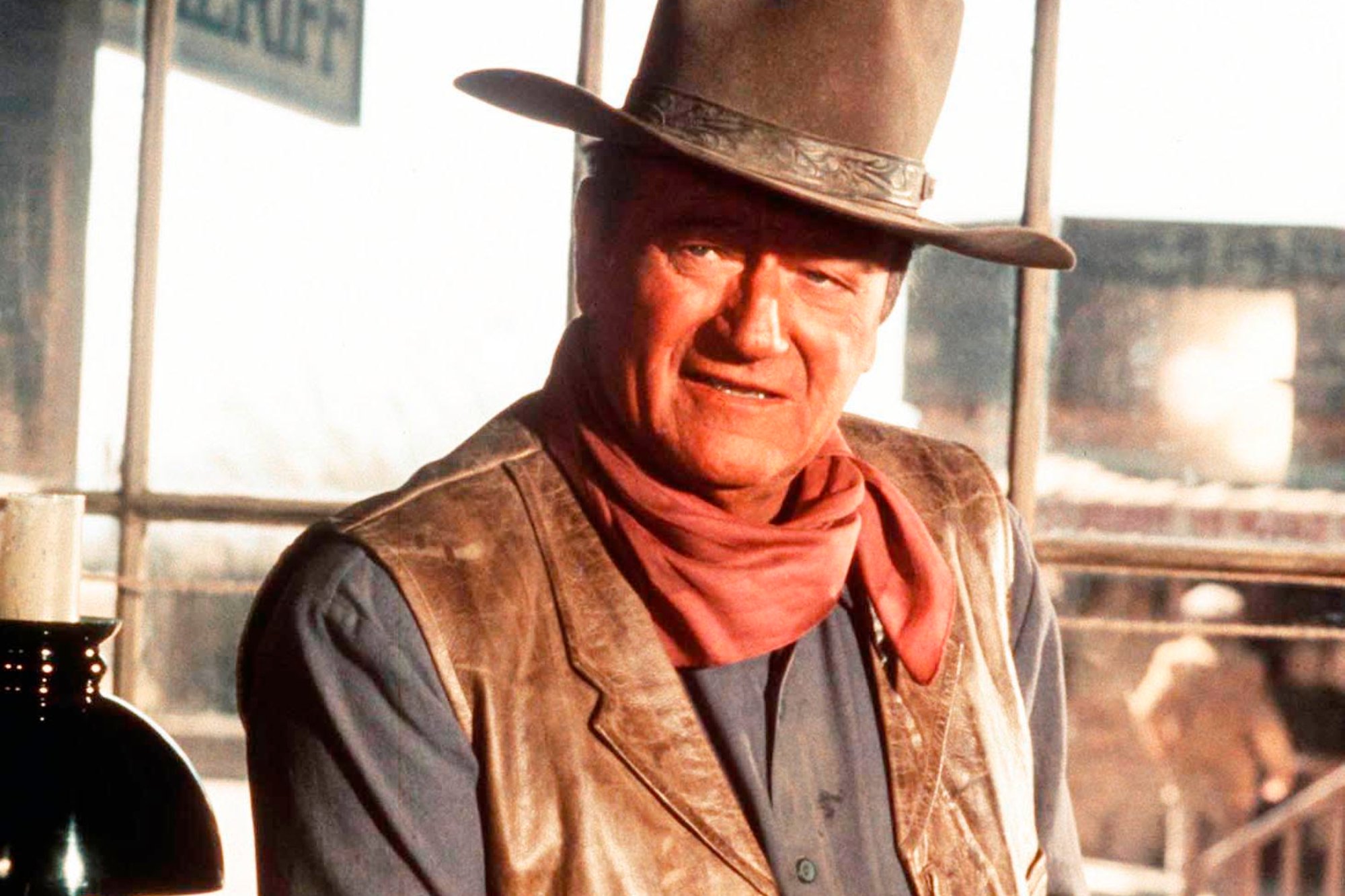 John Wayne, who had an iconic walk. He has a serious look on his face, wearing a Western cowboy outfit with a scarf, leather vest, and hat.