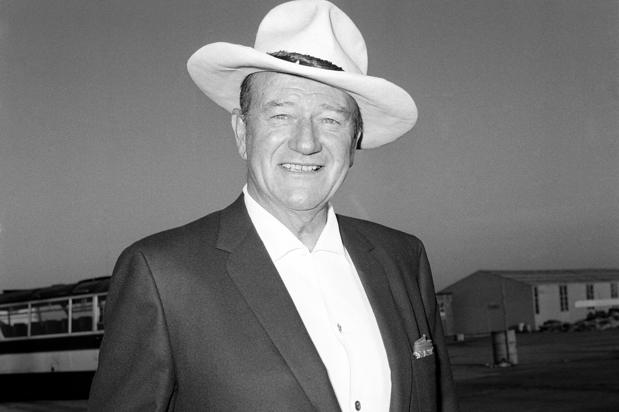 John Wayne, who starred in Western movies. He's smiling wearing a suit jacket and white dress shirt. He has a hat on his head.