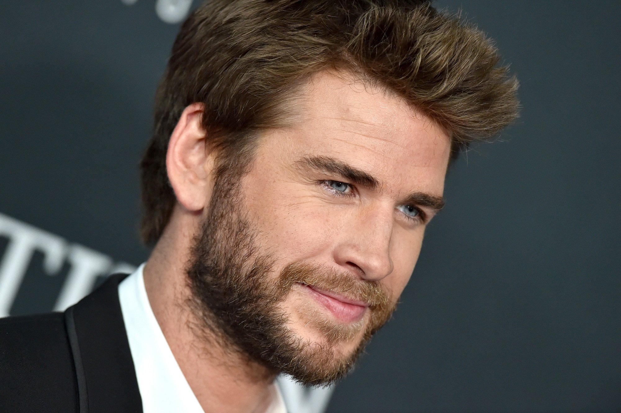 A close-up photo of actor Liam Hemsworth standing in front of a black background with white writing