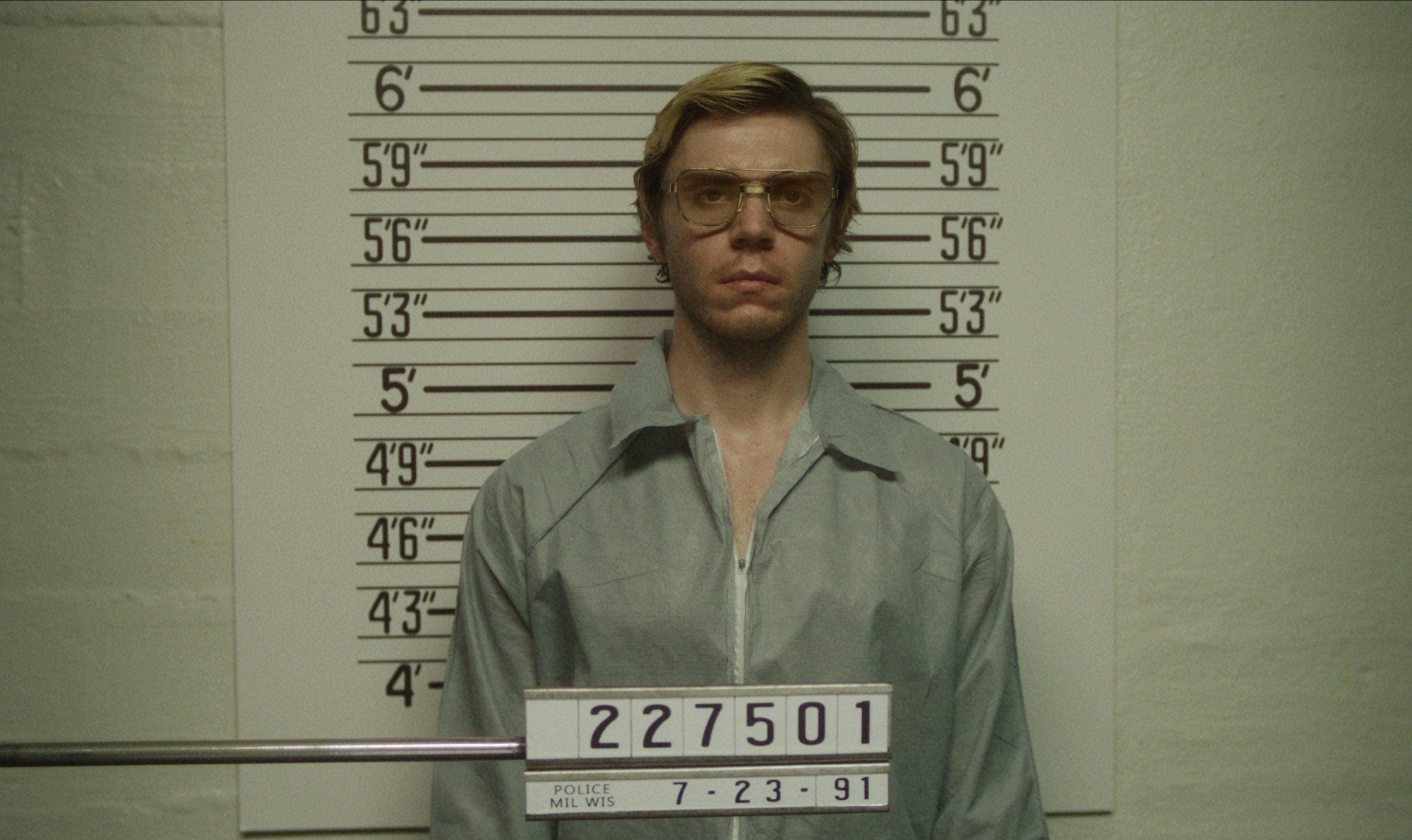 Evan Peters as Jeffrey Dahmer in 'Monster: The Jeffrey Dahmer Story' for our article about its initial ratings. He's taking a prison photo wearing glasses and a grey collared shirt.