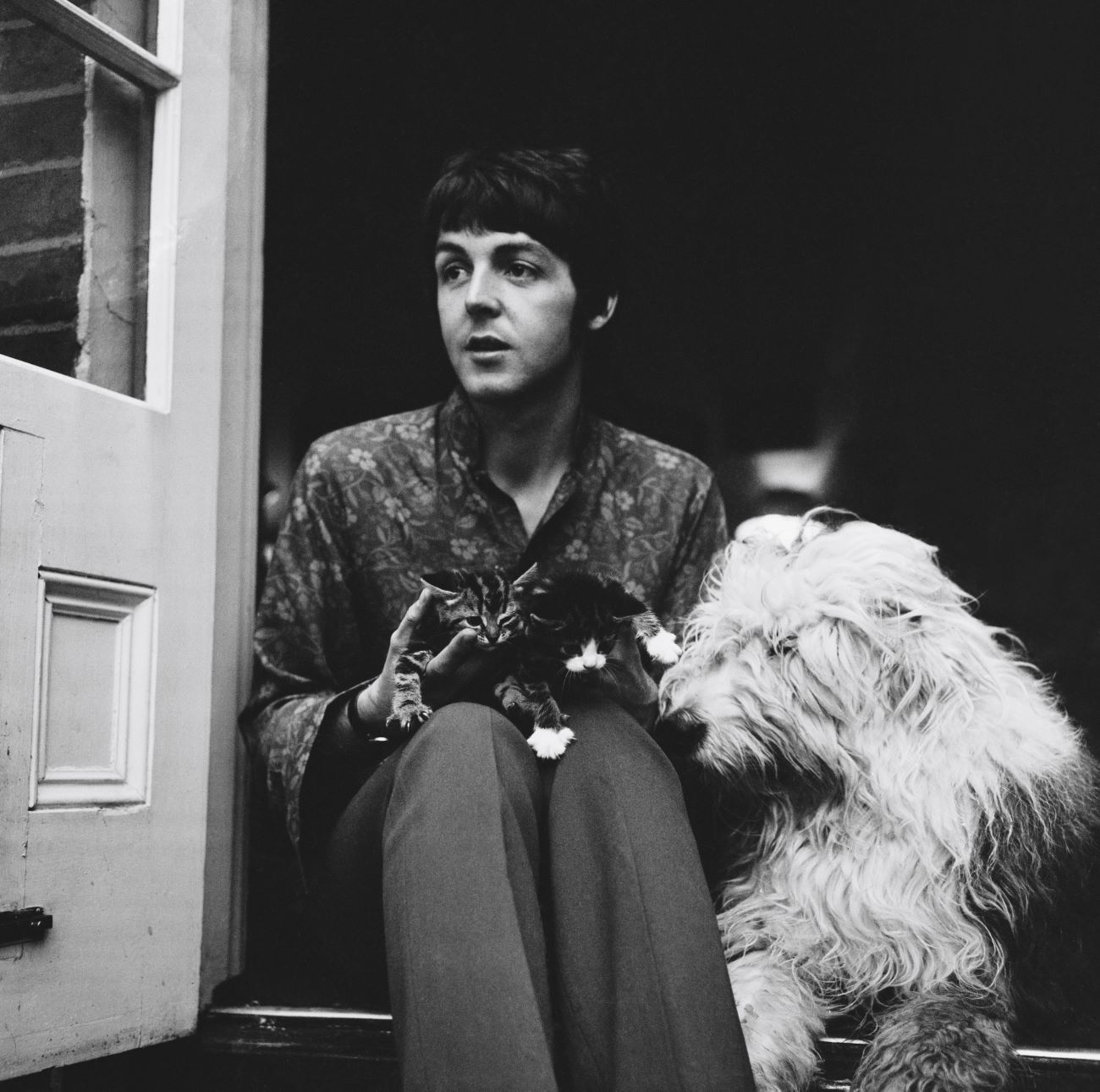 A black and white picture of Paul McCartney sitting in a doorway with a dog and kittens on his lap.