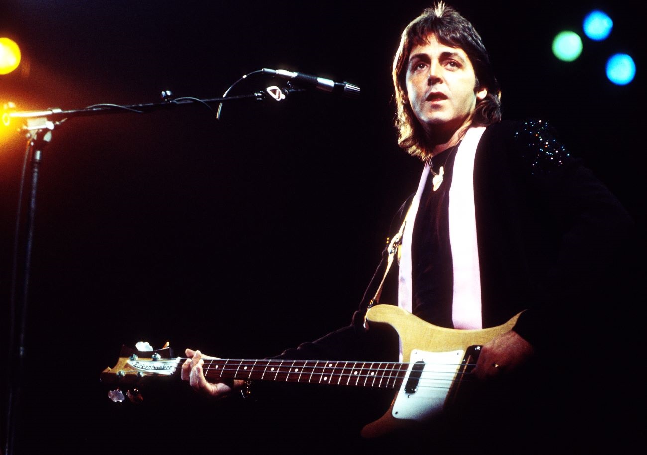 Paul McCartney stands in front of a microphone with a guitar.