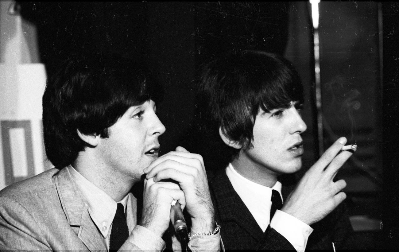 A black and white photo of Paul McCartney and George Harrison sitting next to each other. McCartney holds a microphone and Harrison holds a cigarette.