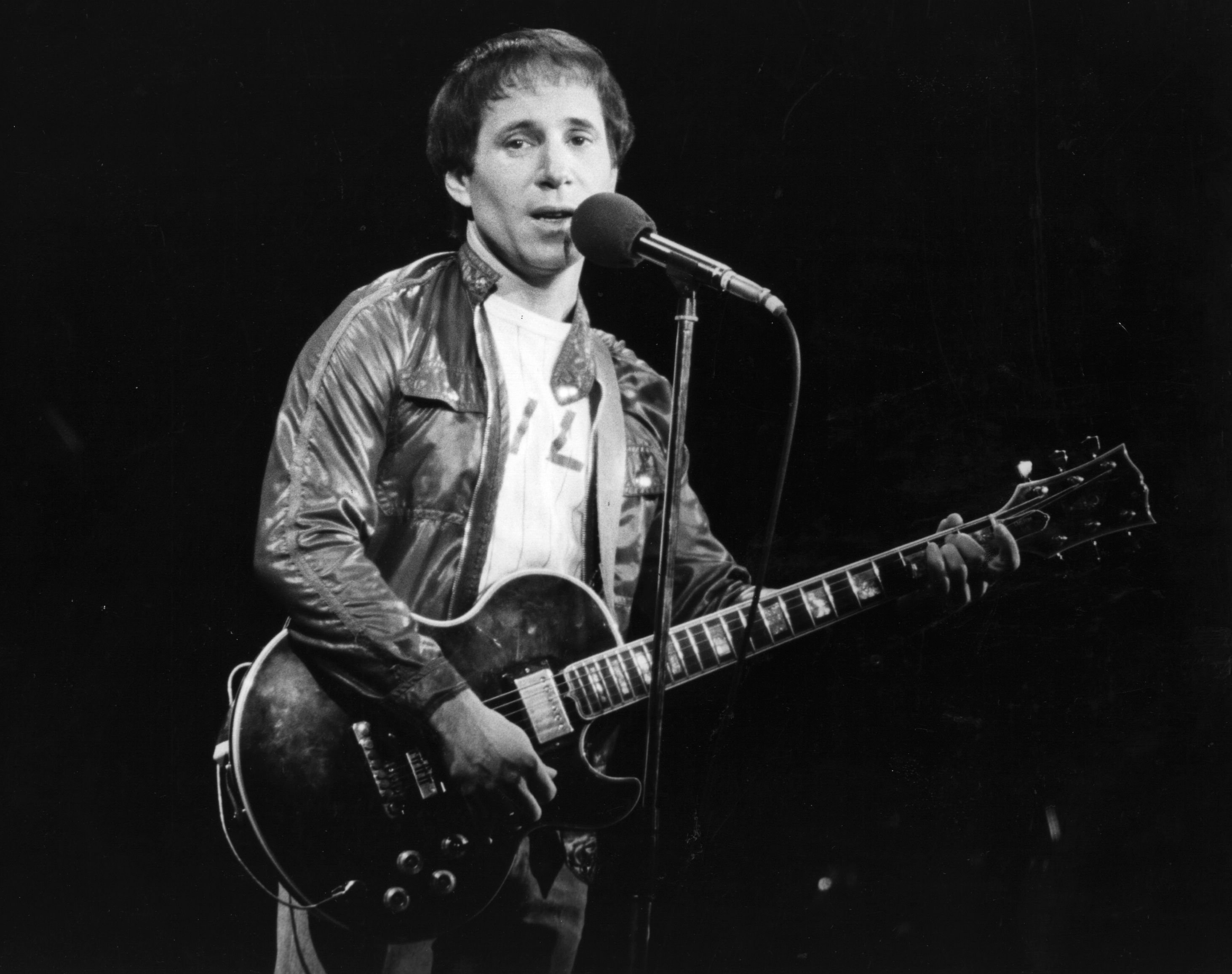 A black and white picture of Paul Simon playing guitar and singing into a microphone.