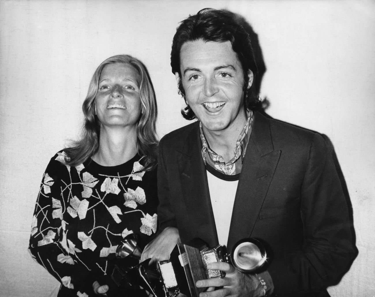 A black and white picture of Linda McCartney standing arm in arm with Paul McCartney, who holds a camera.