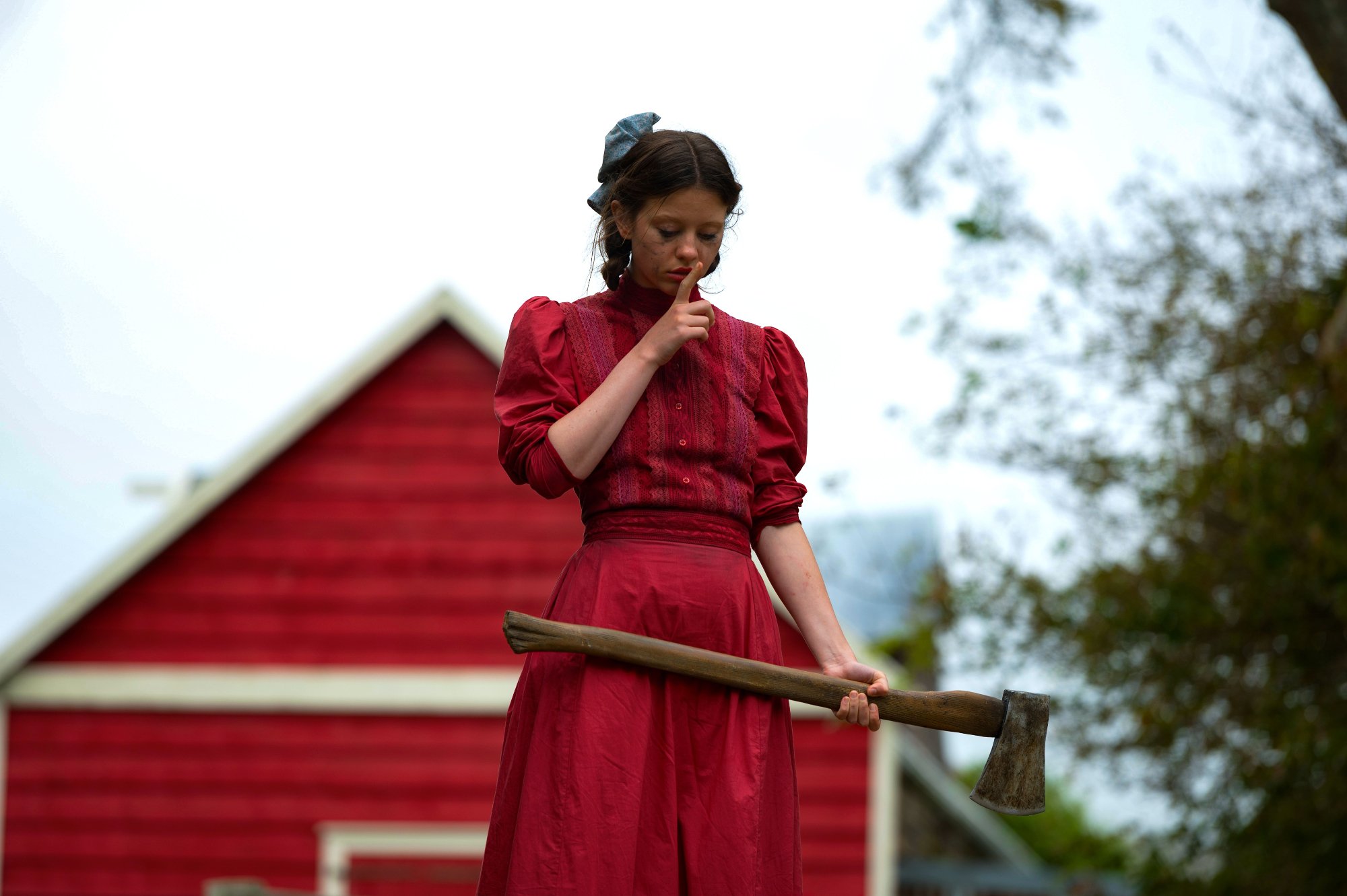 'Pearl' Mia Goth as Pearl wearing a red dress, holding an ax in front of a barn. She's holding a finger up to her lips to shush someone.