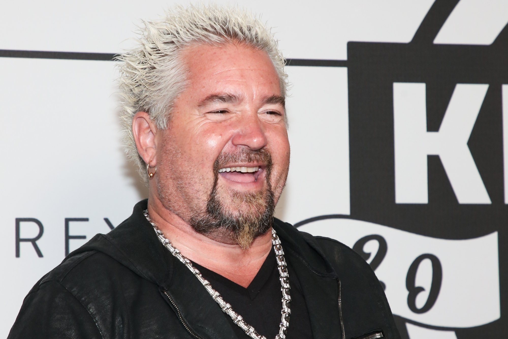 Potential future 'Saturday Night Live' host Guy Fieri. He's giving an open mouth smile, wearing a black shirt, and a chain necklace in front of a backdrop