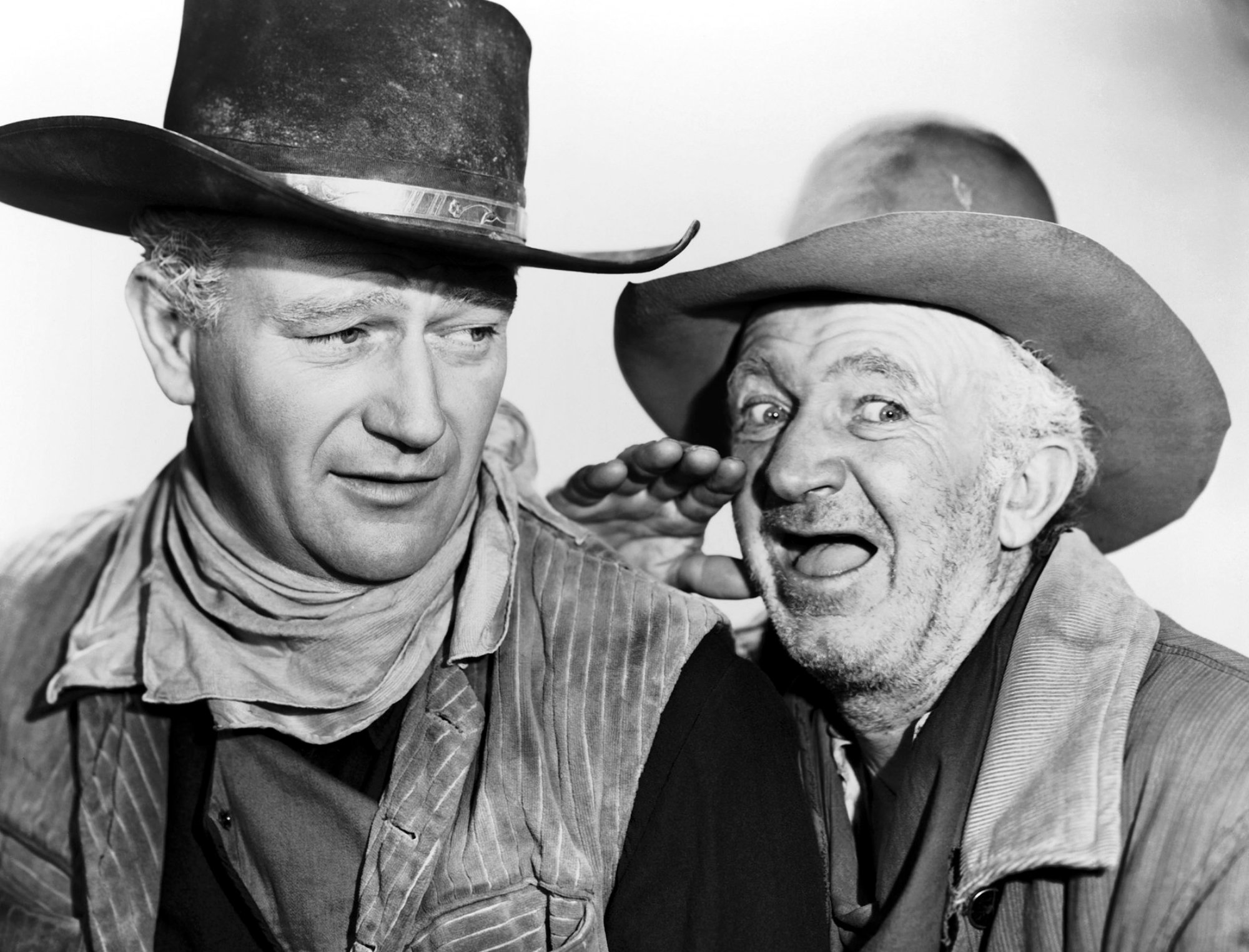 John Wayne and Walter Brennan. Black-and-white photograph of Brennan whispering into Wayne's ear. They're both wearing Western outfits with cowboy hats.