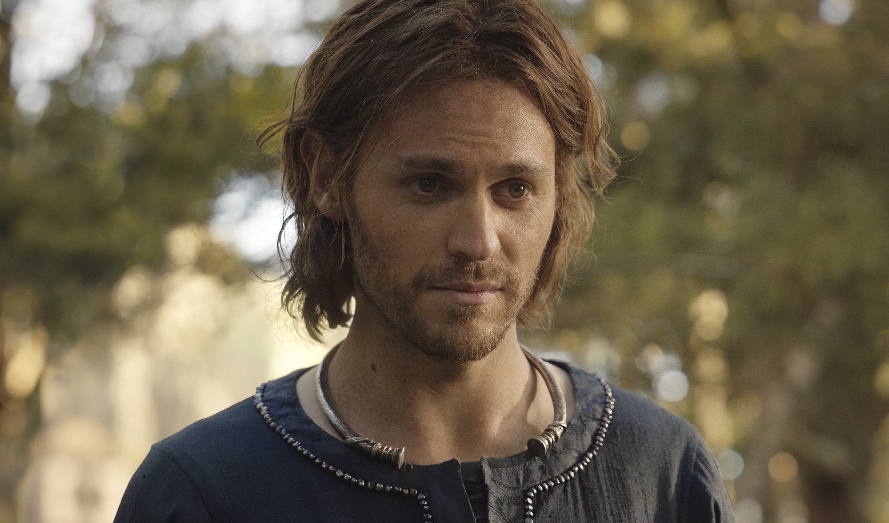 Charlie Vickers as Halbrand, who is revealed to be Sauron in 'The Rings of Power.' He's wearing a blu shirt and smiling.