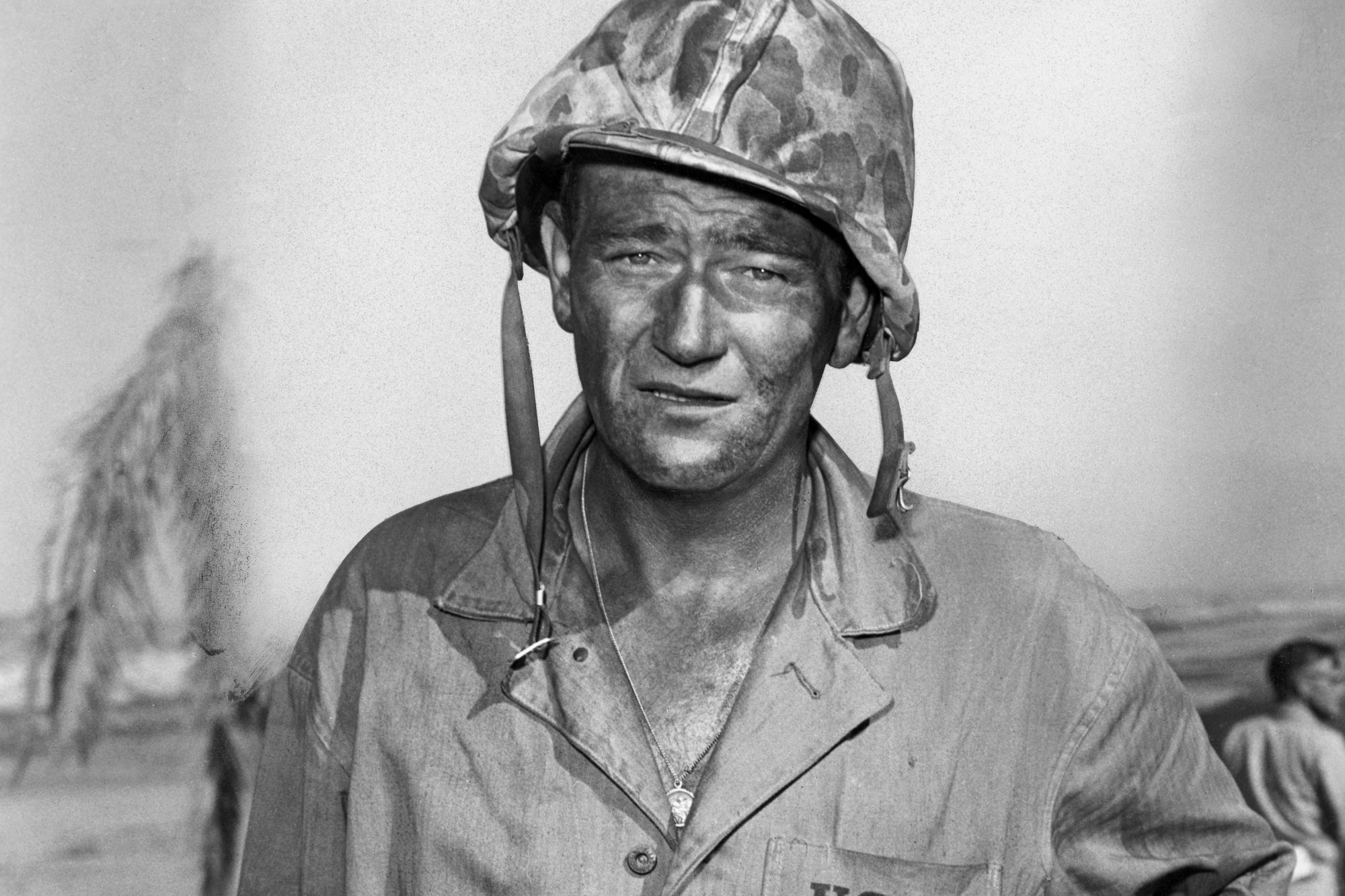 'Sands of Iwo Jima' star John Wayne with a serious look toward the camera in a black-and-white photograph. He's wearing a military uniform and helmet.