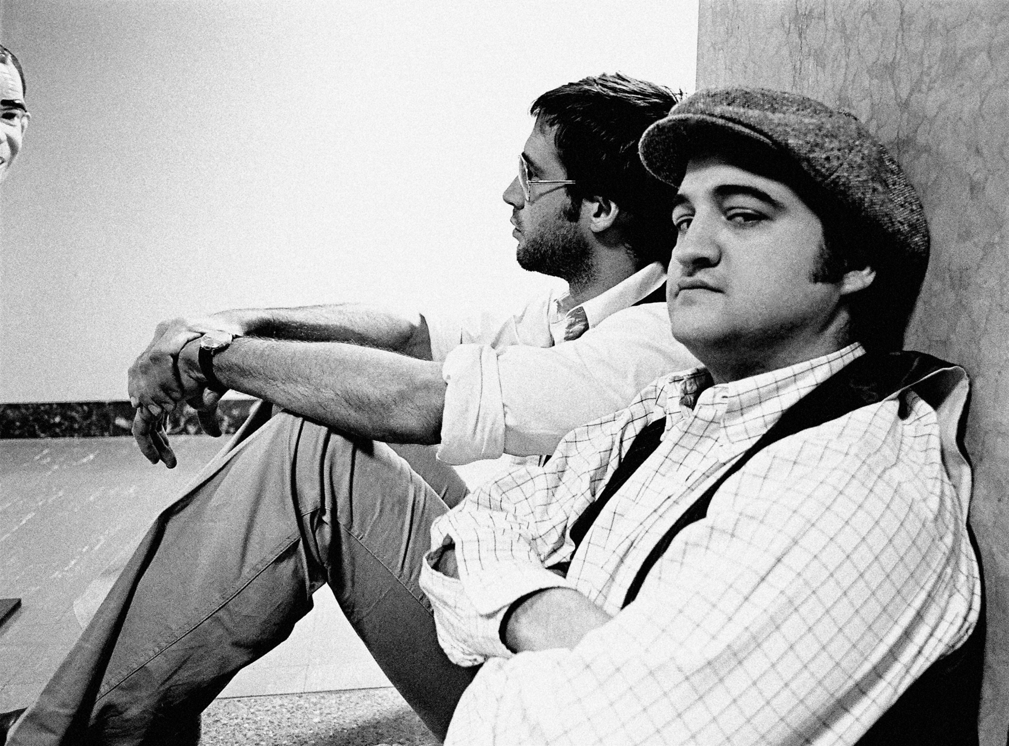 'Saturday Night Live' Chevy Chase and John Belushi sitting against a wall in a black-and-white picture. Chase is looking off and Belushi is looking toward the camera.