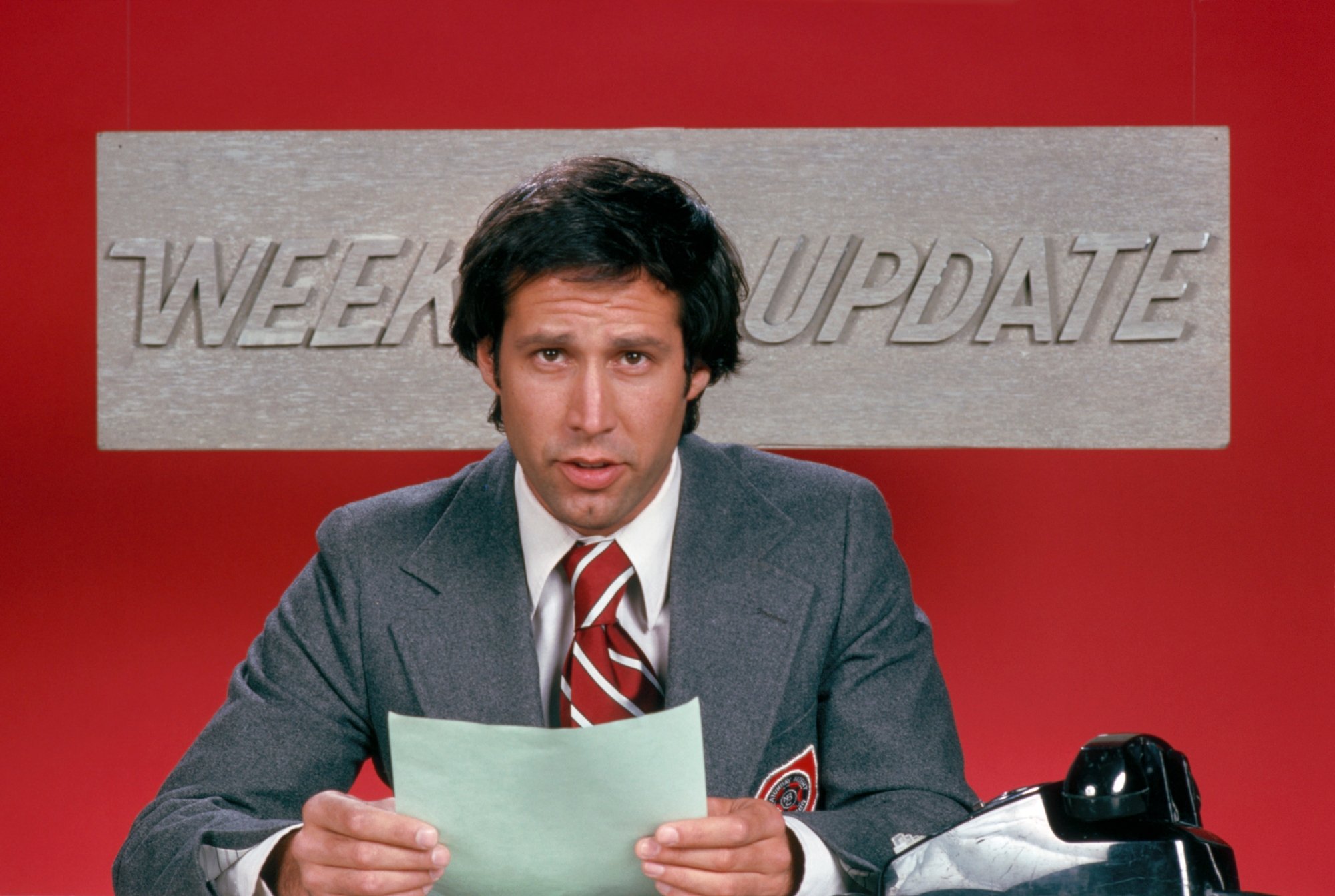 'Saturday Night Live' Chevy Chase on 'Weekend Update' wearing a suit and tie while holding a piece of paper. He's sitting in front of a red background with the 'Weekend Update' logo behind him.