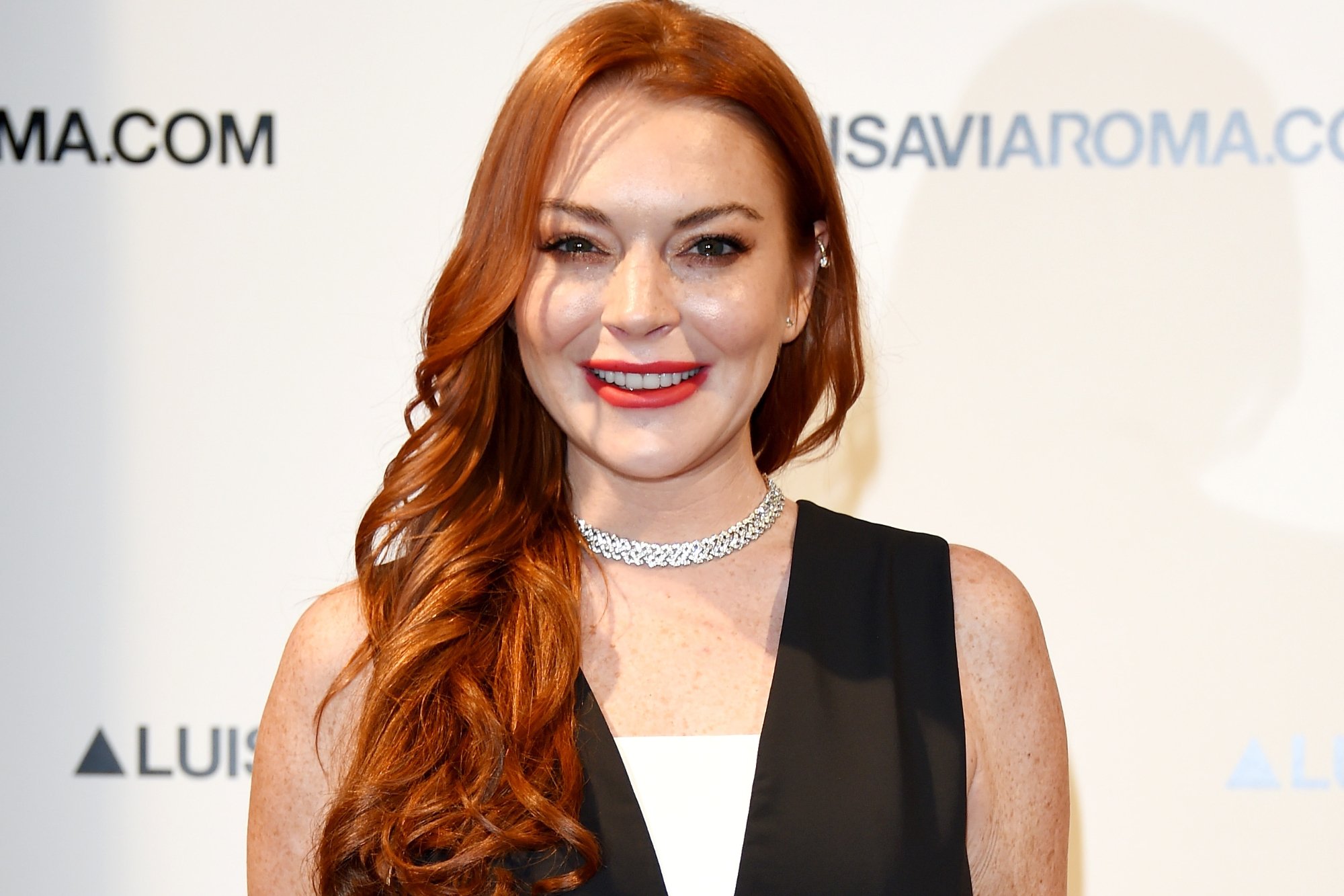 'Saturday Night Live' host Lindsay Lohan wearing a black and white top. She's smiling standing in front of a white step-and-repeat.