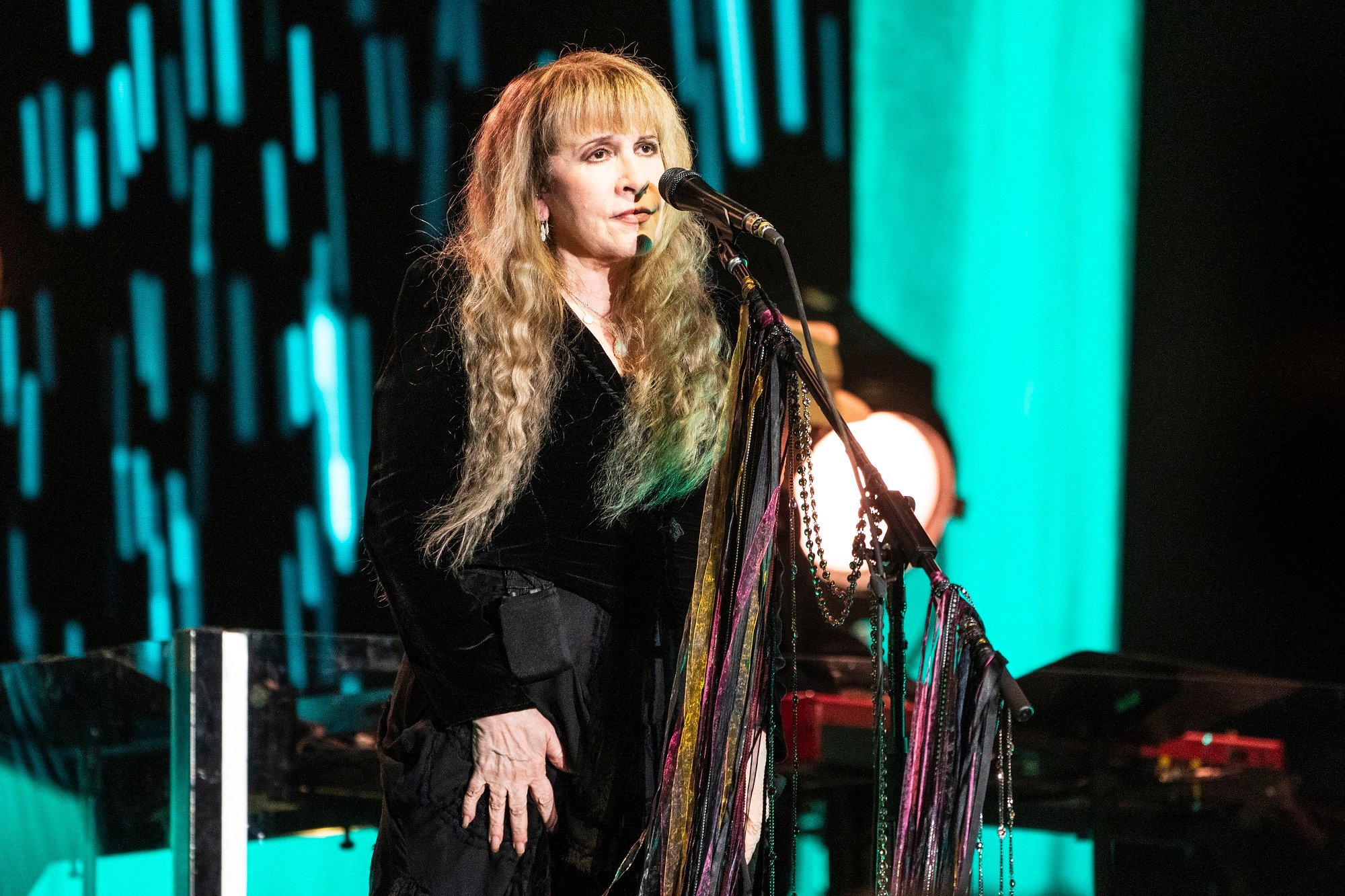 Stevie Nicks stands on stage in front of a mic stand while wearing a long-sleeved black dress