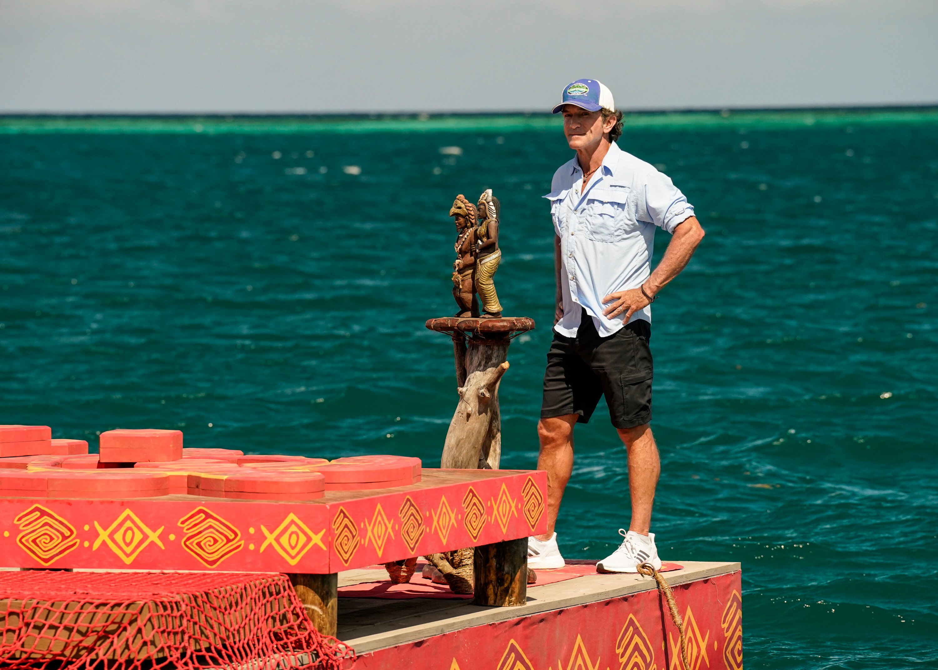 Jeff Probst, the host of 'Survivor' Season 43 on CBS, wears a light blue button-up shirt, black shorts, and a blue and white 'Survivor' baseball cap while standing on a platform in the water.
