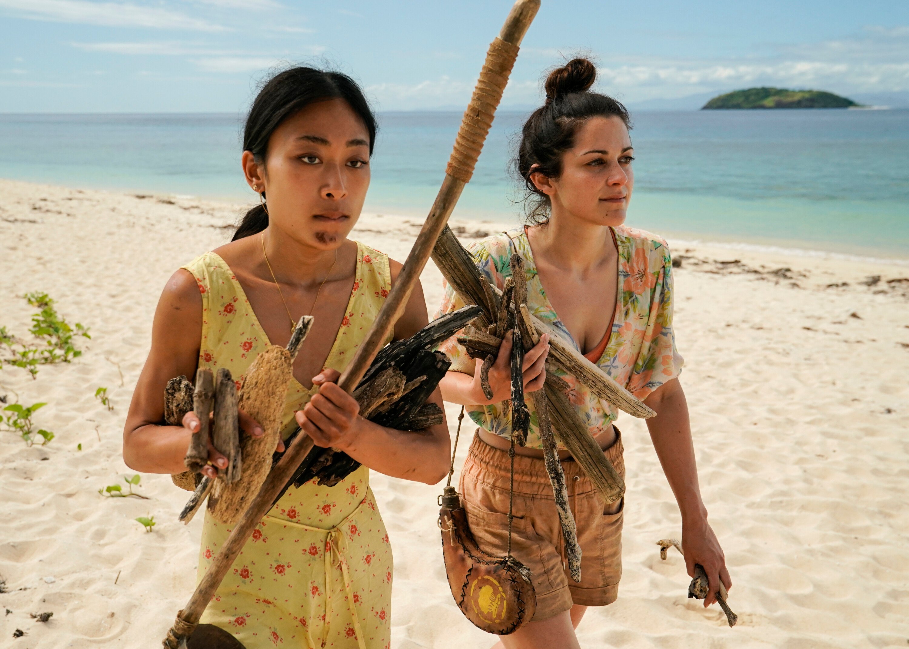 Elie Scott, who, according to 'Survivor' Season 43 spoilers, may win, collects firewood with tribemate Jeanine Zheng on the beach. Jeanine wears a light yellow romper with red flowers. Elie wears a pink, blue, green, and white floral shirt and muted orange shorts.