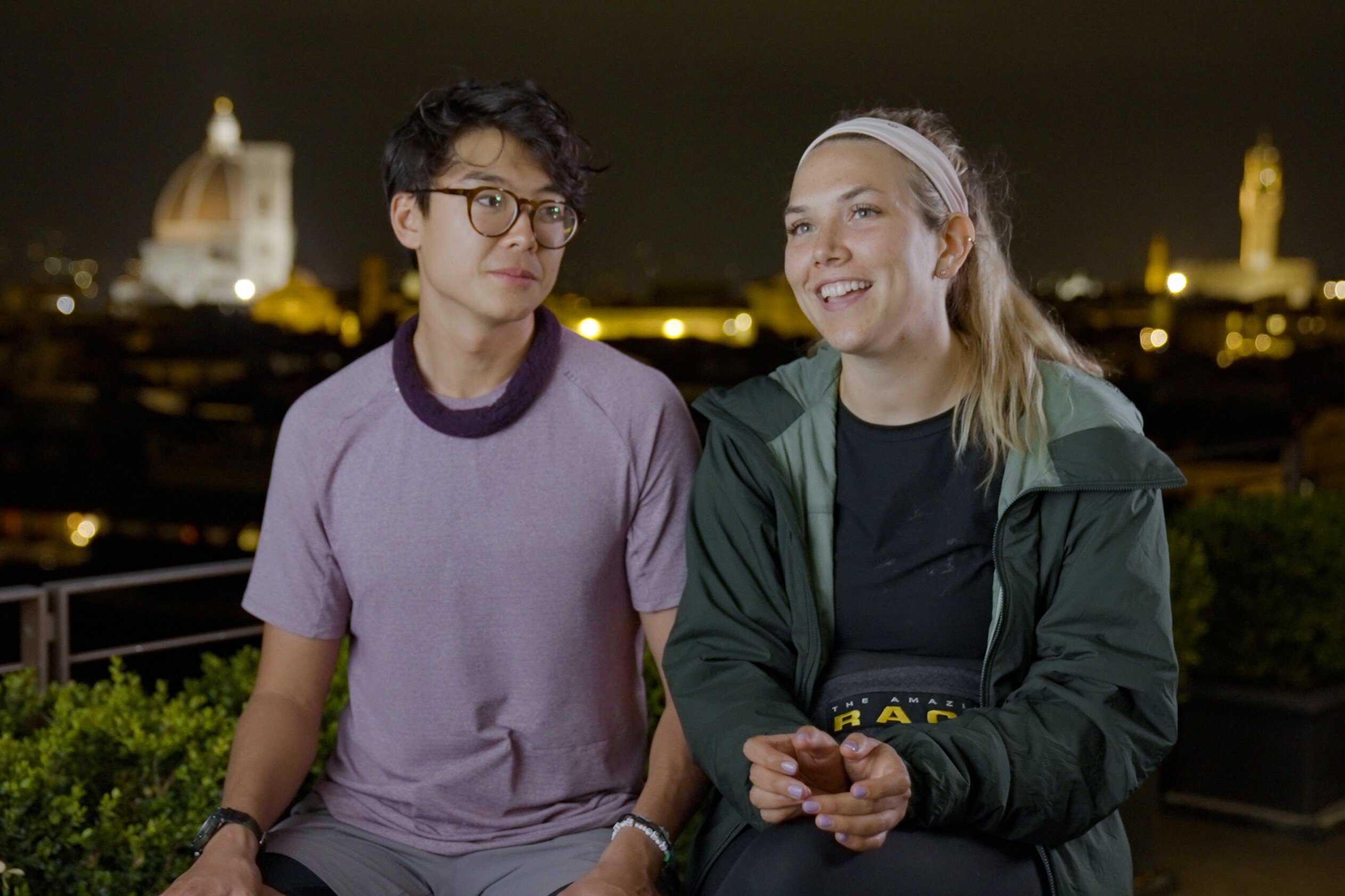 Derek Xiao and Claire Rehfuss, who might be the winner of 'The Amazing Race' Season 34 on CBS, sit next to each other while filming a confessional at night. Derek wears a purple shirt and gray shorts. Claire wears a green jacket over a black shirt and black pants.