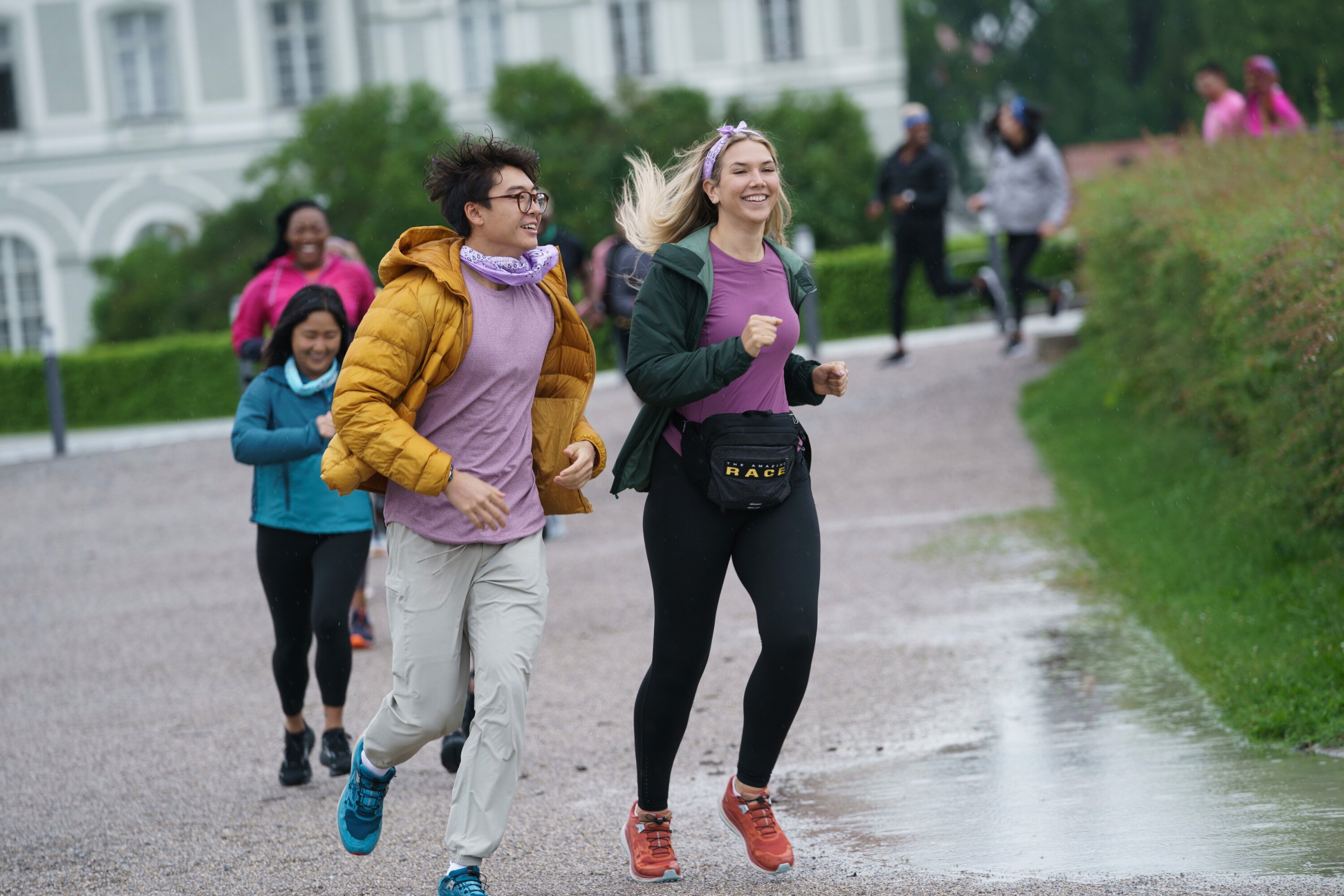 Derek Xiao and Claire Rehfuss, who star in 'The Amazing Race' Season 34 on CBS, run to the starting line. Derek wears a mustard yellow jacket over a light purple shirt and gray sweatpants. Claire wears a dark green jacket over a purple shirt and black leggings.
