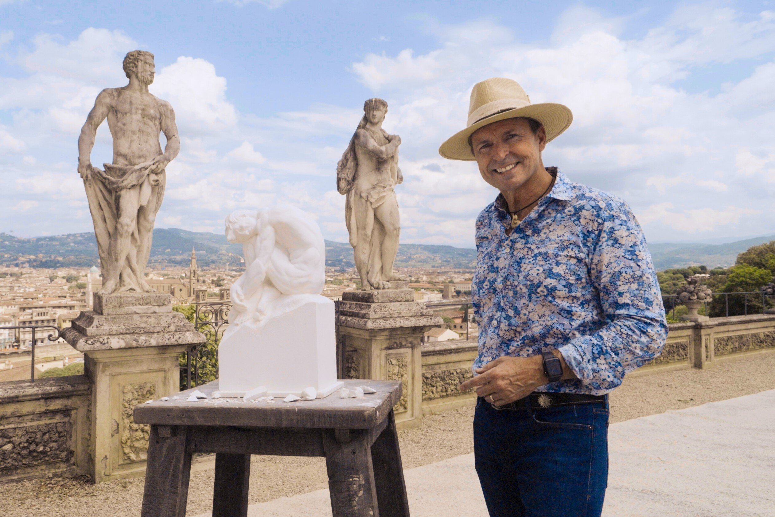Phil Keoghan, who hosts 'The Amazing Race' Season 34 on CBS, wears a blue floral long-sleeved button-up shirt, jeans, and a tan hat while posing in front of statues in Italy.