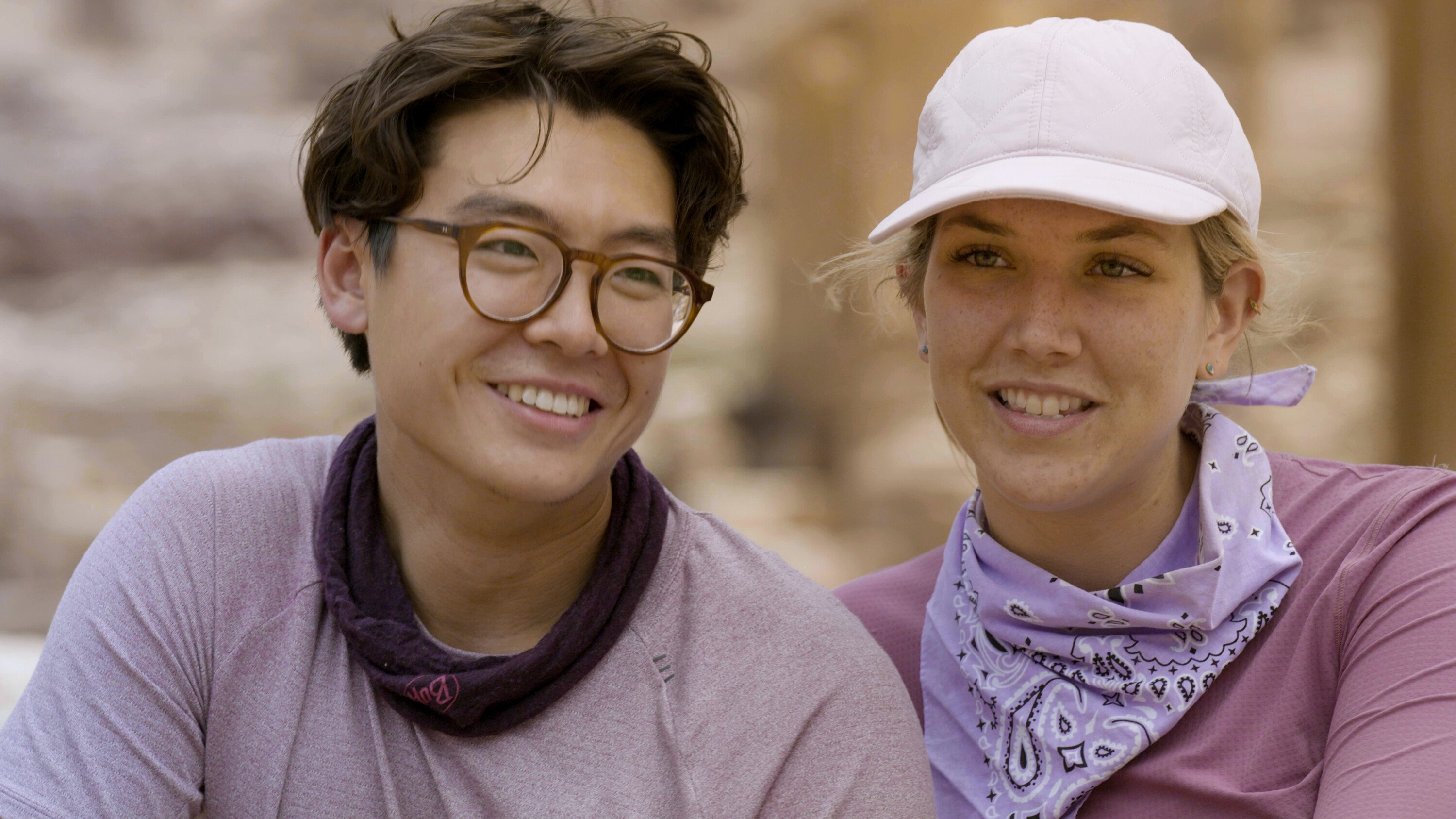 Derek Xiao and Claire Rehfuss, who starred in 'The Amazing Race' Season 34 Episode 5 on CBS, film a confessional. Derek wears a light purple shirt, dark purple band around his neck, and glasses. Claire wears a purple long-sleeved shirt, purple bandana, and light purple baseball cap.