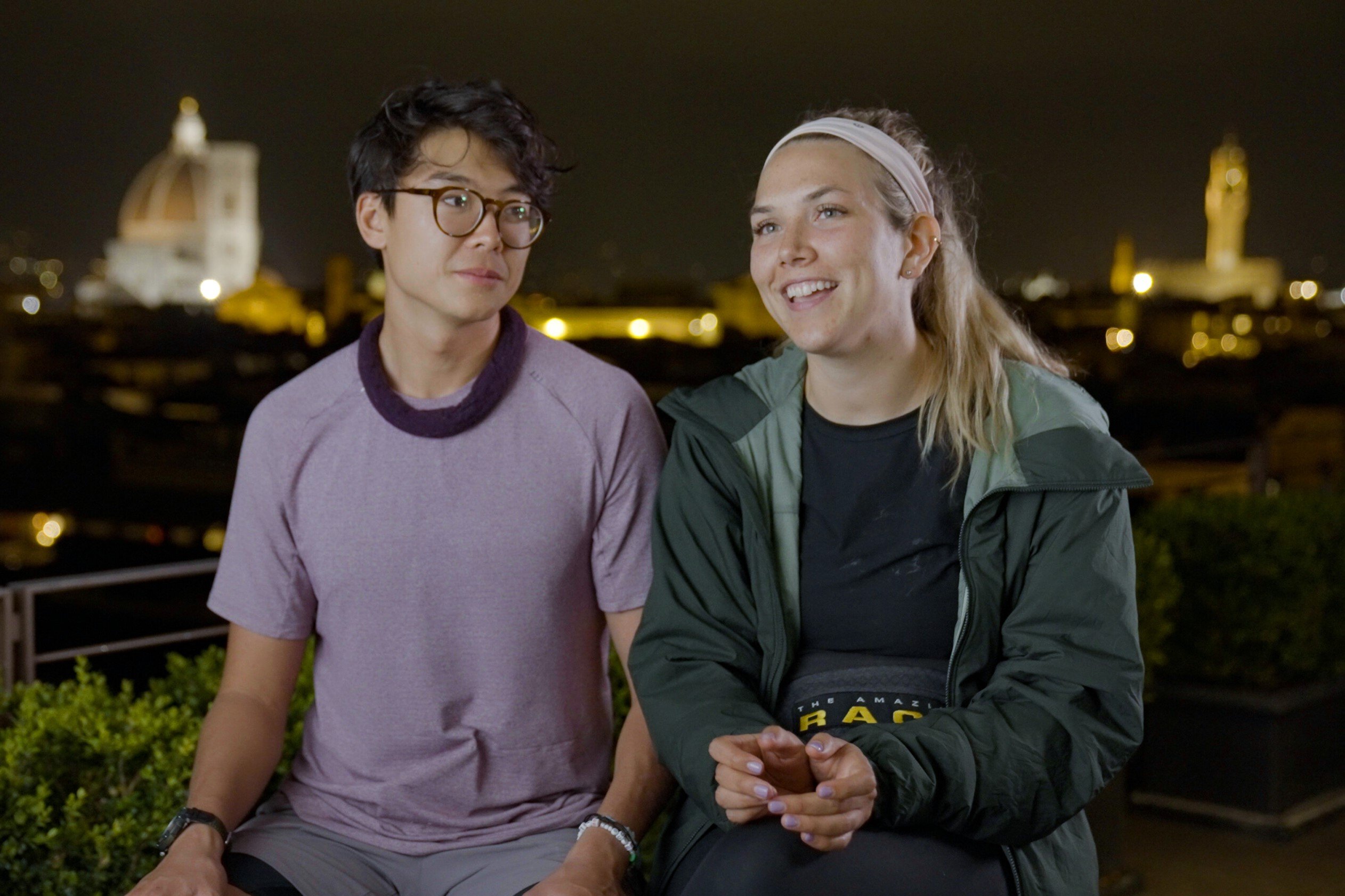 Derek Xiao and Claire Rehfuss, who star in 'The Amazing Race' Season 34 on CBS, record a confessional at night. Derek wears a light purple shirt and light gray pants. Claire wears a dark green jacket over a black shirt and black leggings.