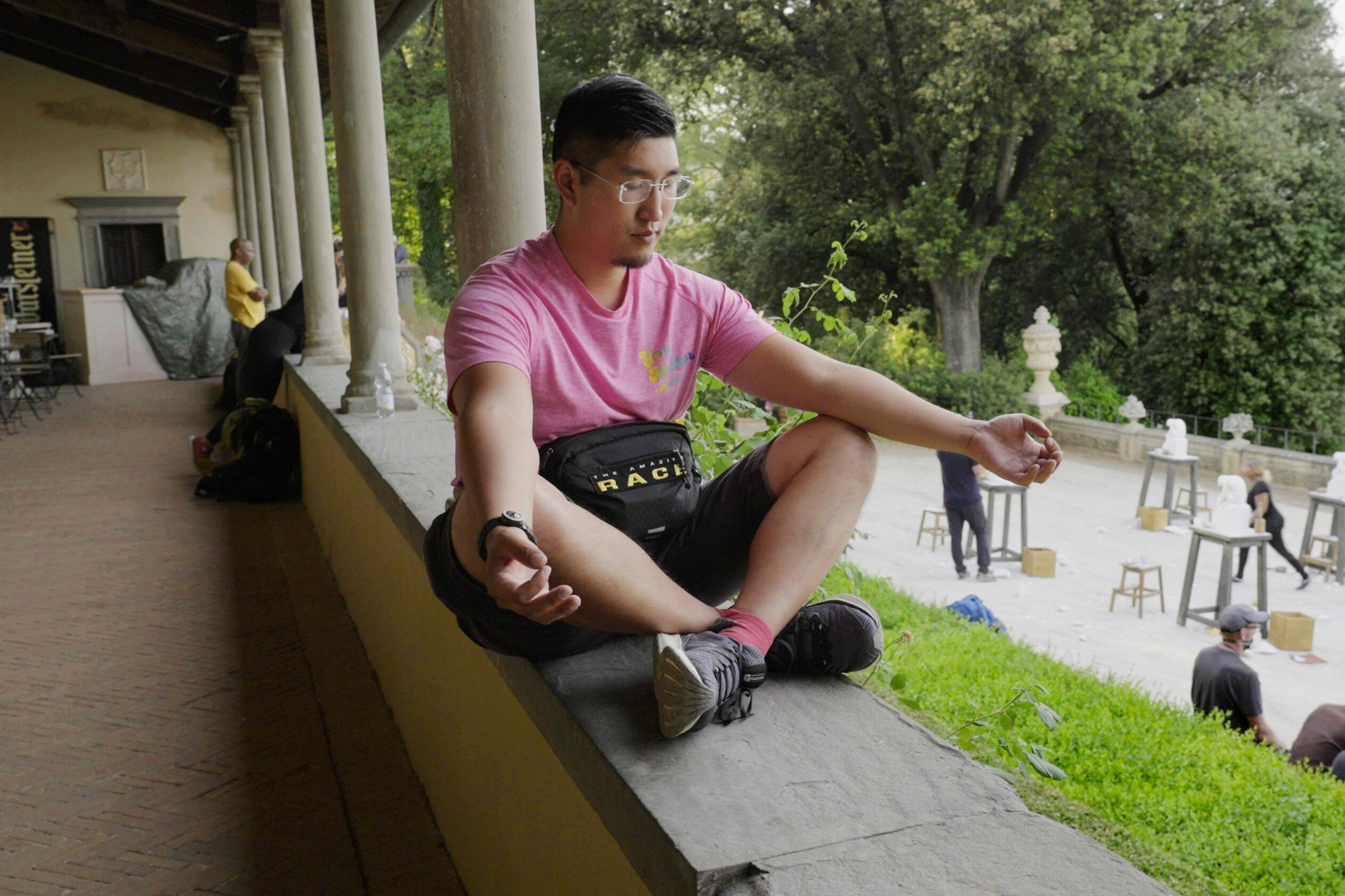 Rich Kuo who stars in 'The Amazing Race' Season 34 Episode 4, which airs tonight, Oct. 12, wears a pink shirt and black shorts while meditating.