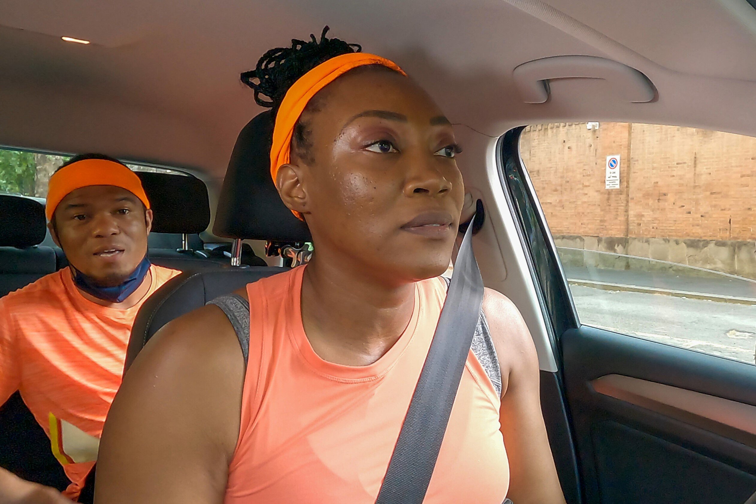 Lumumba Roberts and Glenda Roberts, one of the teams in 'The Amazing Race' Season 34, drive in a car, with Lumumba in the backseat and Glenda in the driver's seat. They both wear orange shirts and orange headbands.