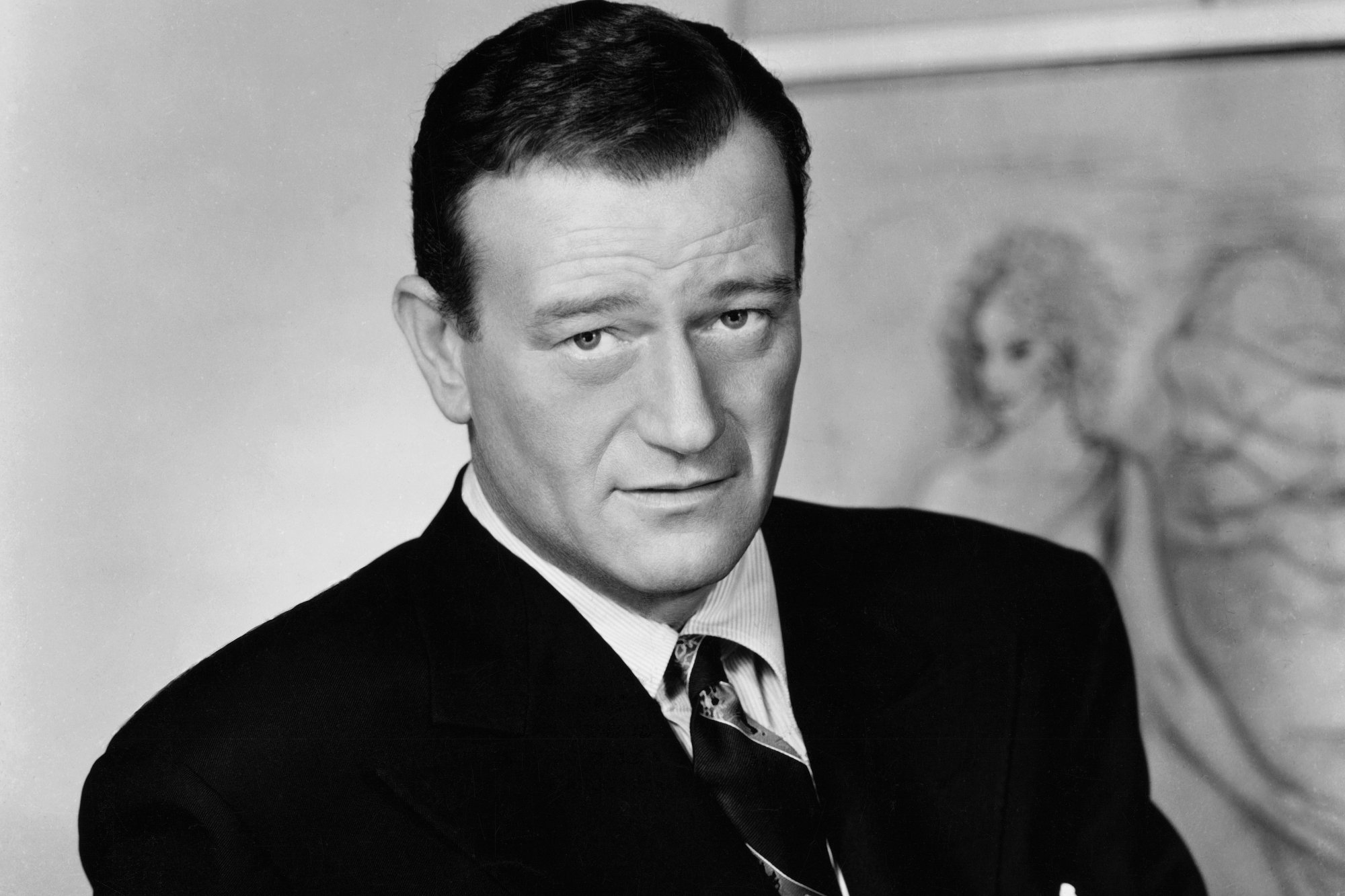 'The Barbarian and the Geisha' star John Wayne wearing a suit and tie, looking directly into the camera in a black-and-white photograph