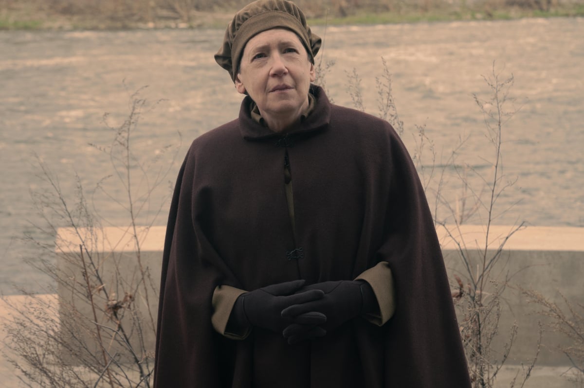 Ann Dowd as Aunt Lydia in The Handmaid's Tale Season 5. Aunt Lydia wears a brown cloak and hat and stands outside in front of a river. 