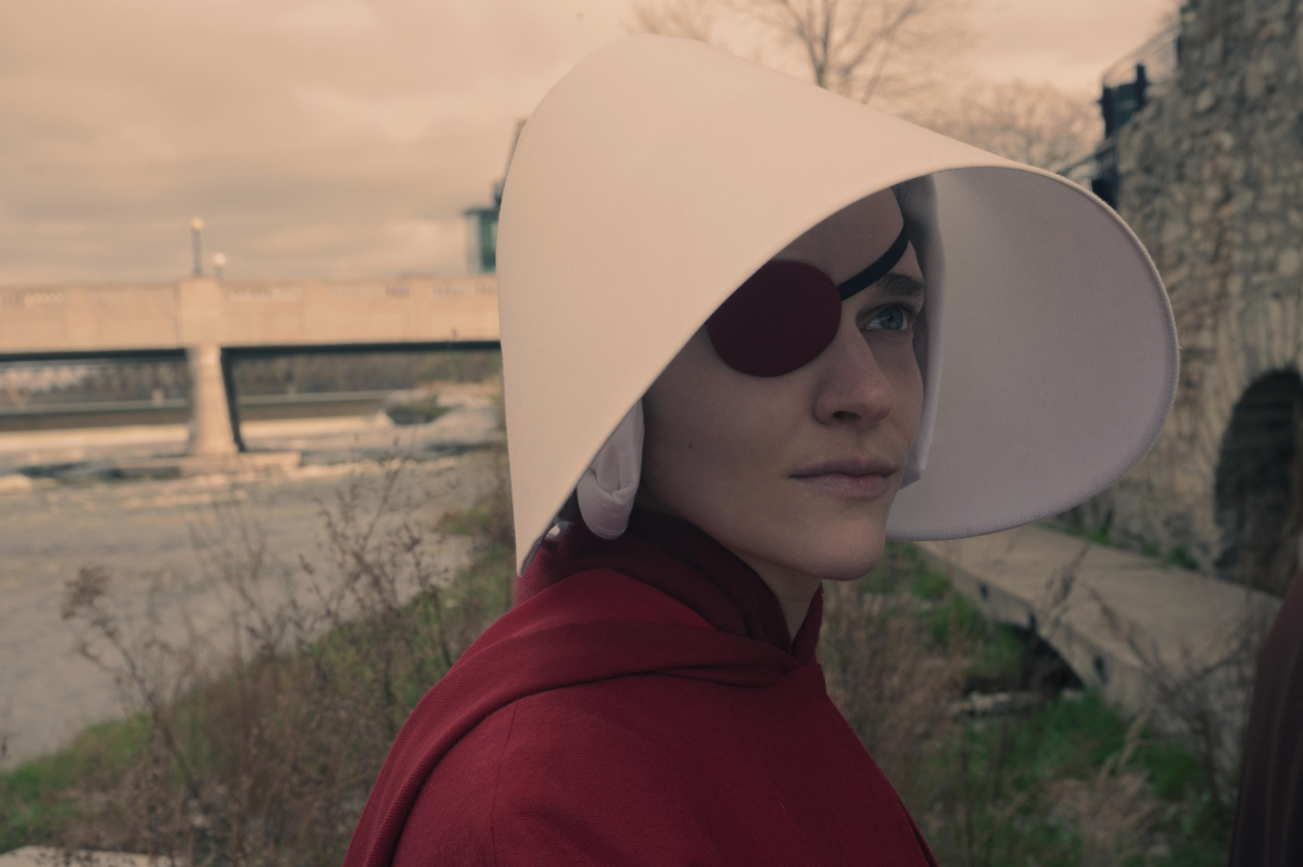 Madeline Brewer as Janine in The Handmaid's Tale Season 5. Janine wears her red cloak and white handmaid wings and stands with a river behind her.