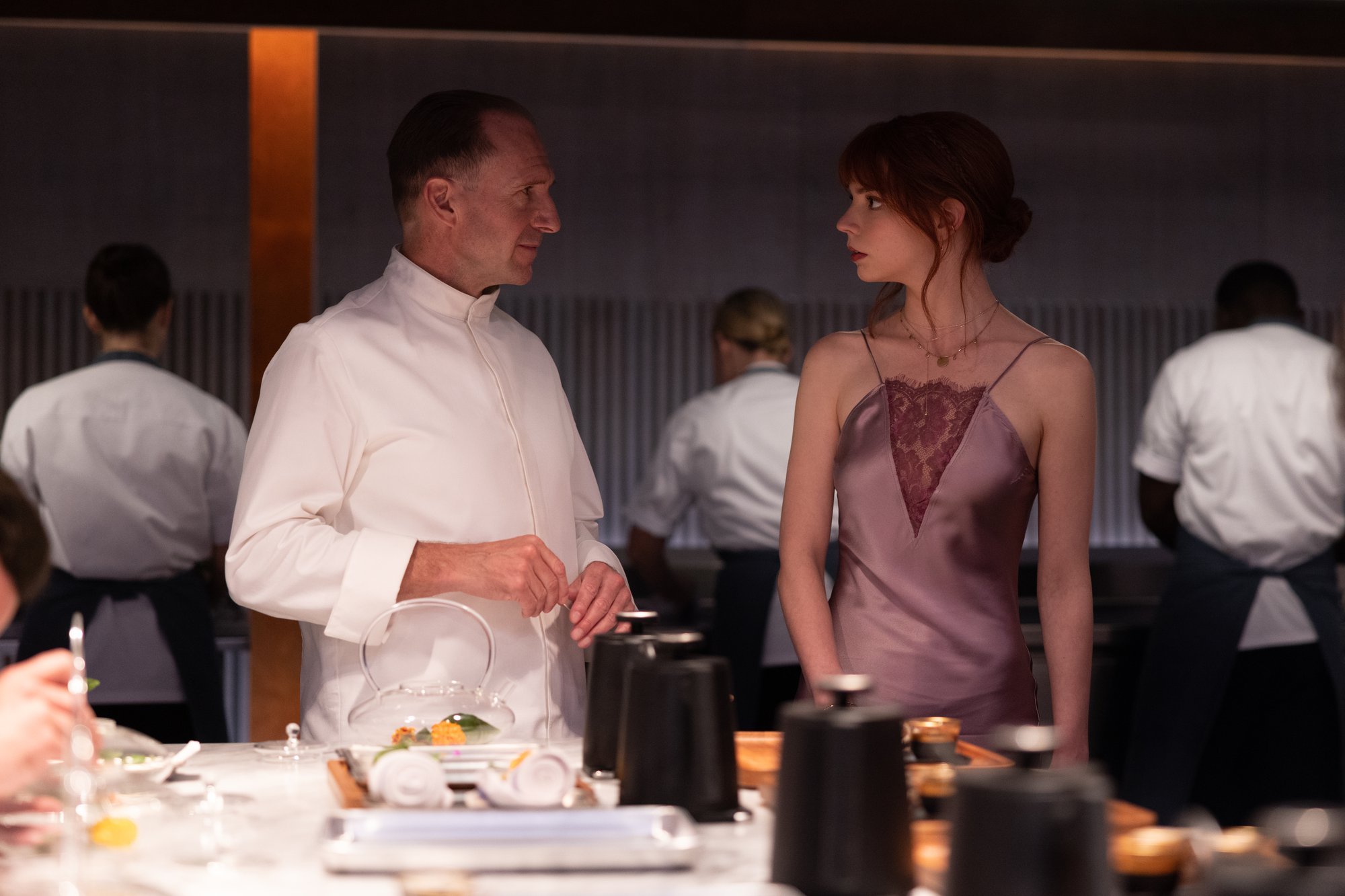 'The Menu' Ralph Fiennes as Chef Slowik and Anya Taylor-Joy as Margot staring at one another in front of a kitchen countertop. Cooks in the background cooking.