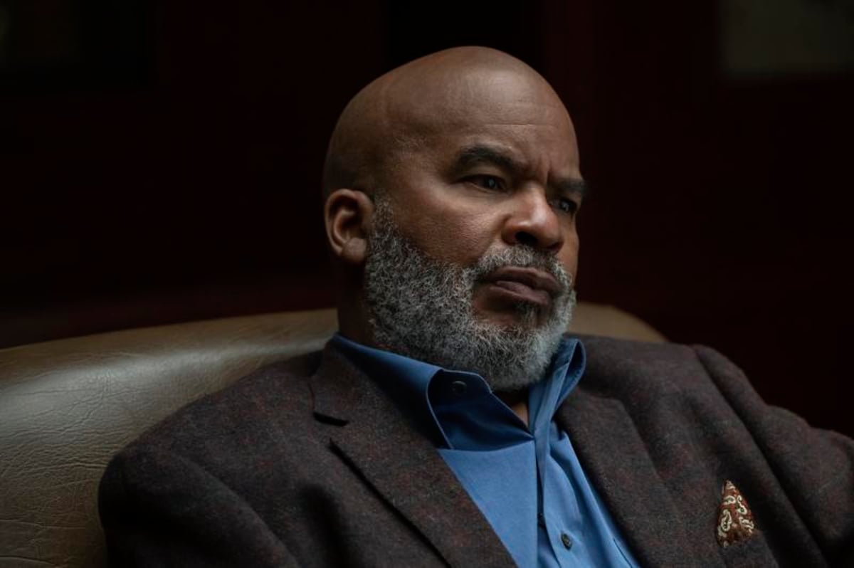 David Alan Grier as Charlie in The Patient. Charlie sits in a chair wearing a brown jacket and blue button-down.