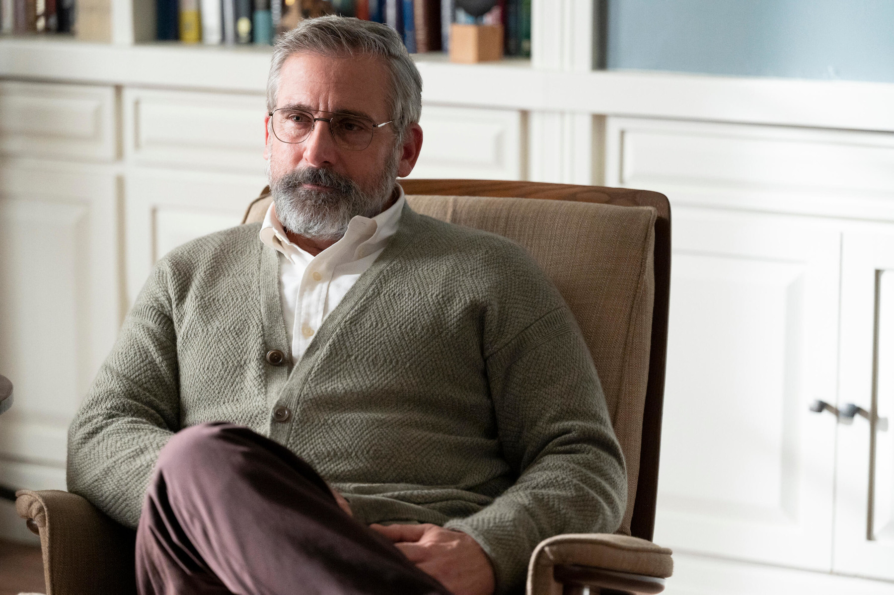 Steve Carell in 'The Patient' for our article about episode 9's release date. He's wearing a grey sweater, sitting in a brown chair, and crossing one leg over the other.