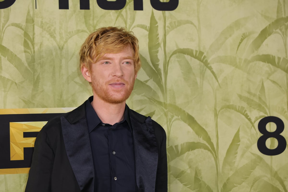 The Patient star Domhnall Gleeson wears a black suit jacket and button-down and stands in front of a green backdrop. 