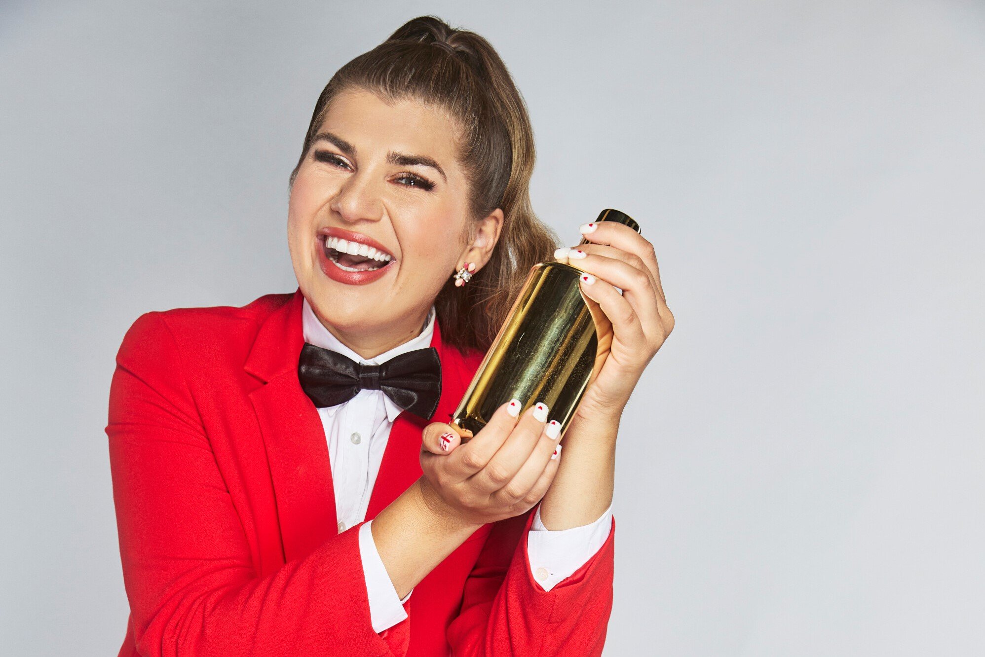 Ezra Freeman, who stars as the bartender in 'The Real Love Boat' on CBS, wears a red suit over a white button-up shirt and black bow tie while holding a drink shaker.