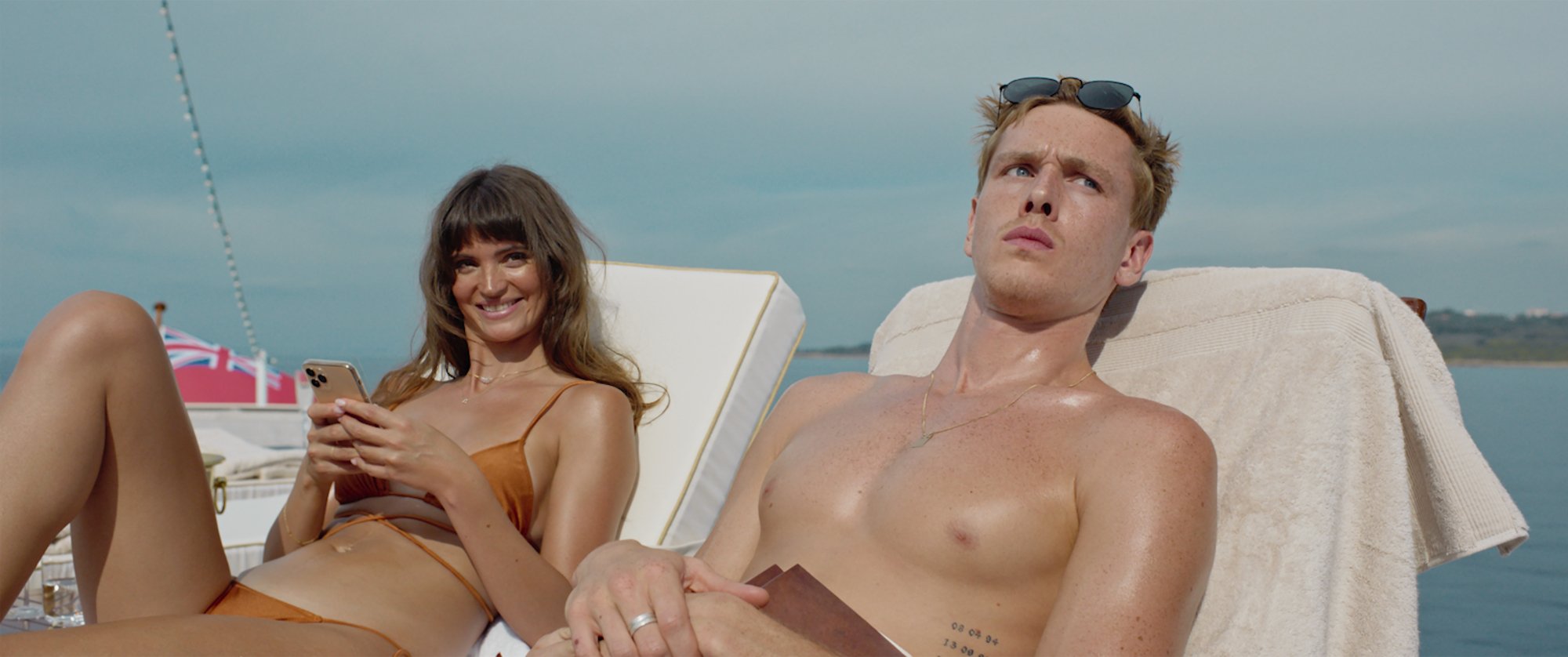 'Triangle of Sadness' Charlbi Dean as Yaya and Harris Dickinson as Carl sitting on lounge chairs in their swimwear. Yaya is on her iPhone.