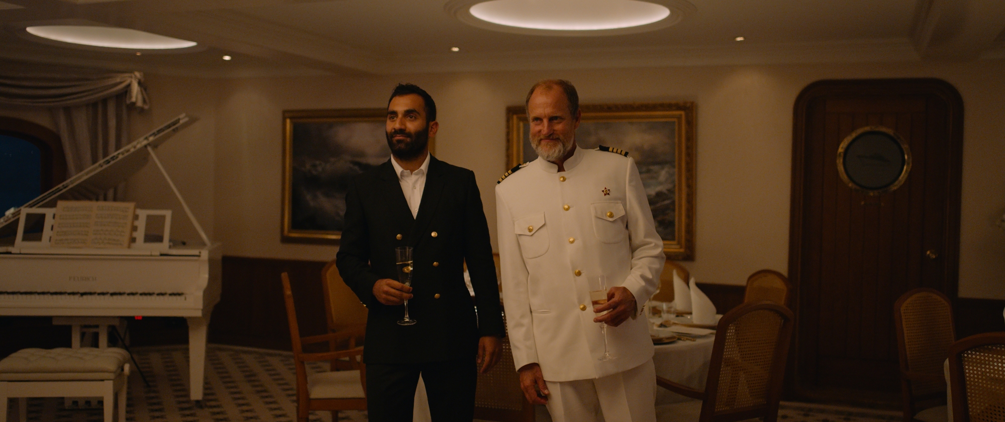 'Triangle of Sadness' Darius as Arvin Kananian and Woody Harrelson as The Captain with close-mouthed grins. They're wearing formal crew and captain-wear while holding a glass of champagne.