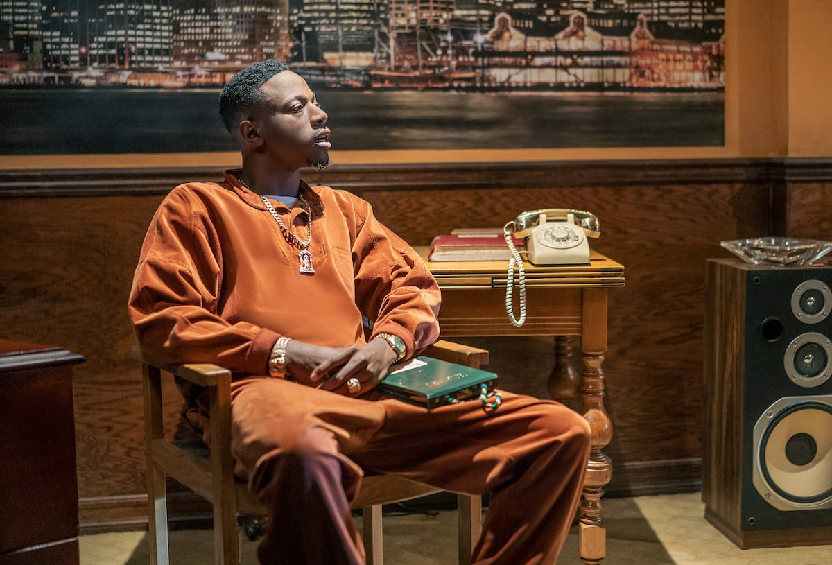 Joey Bada$$ as Unique wearing an orange outfit sitting in a chair in 'Power Book III: Raising Kanan'