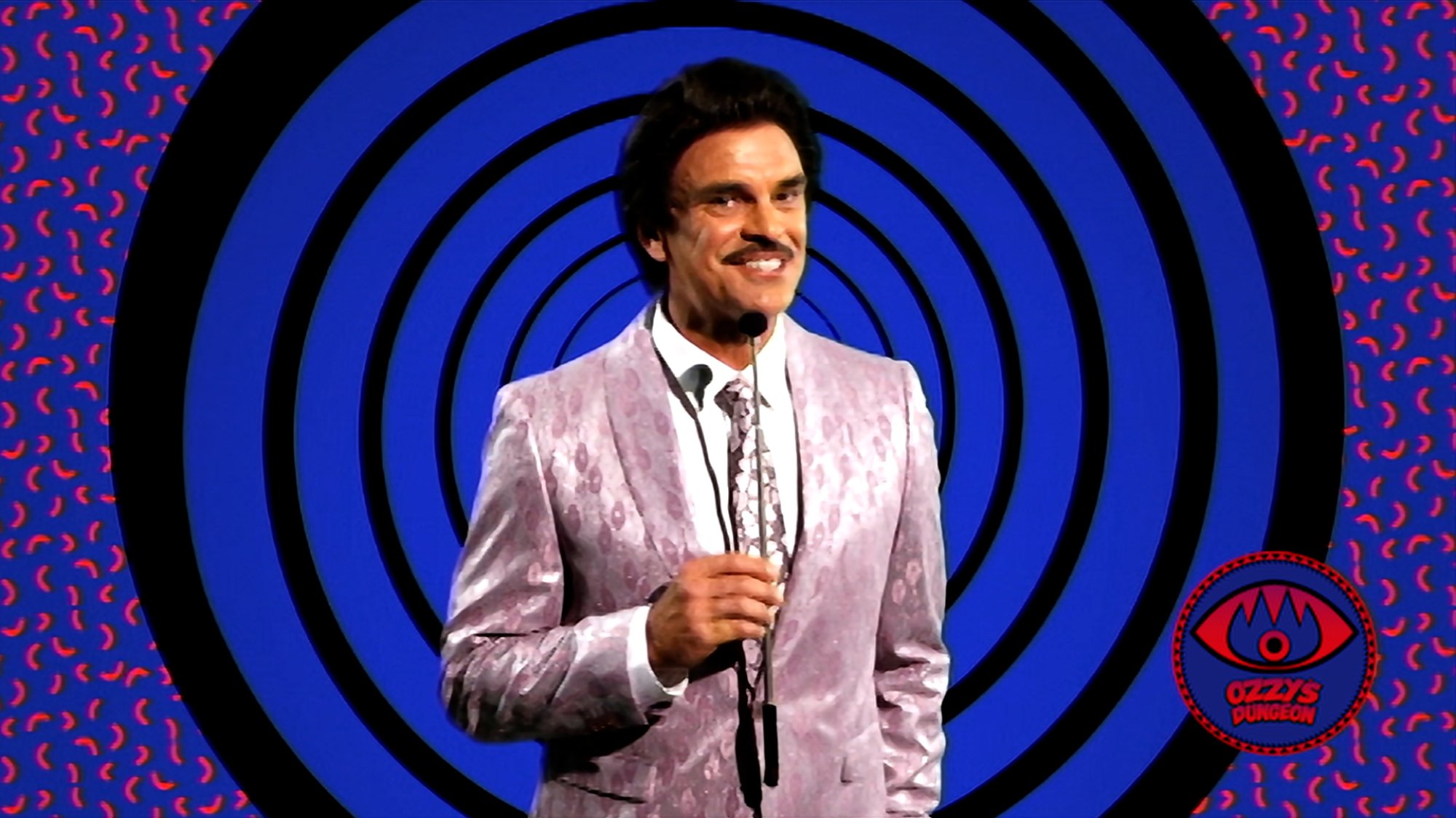 'V_H_S_99' Steven Ogg as Host wearing a pink suit with a white collared dress shirt, holding a microphone in front of a swirly blue background