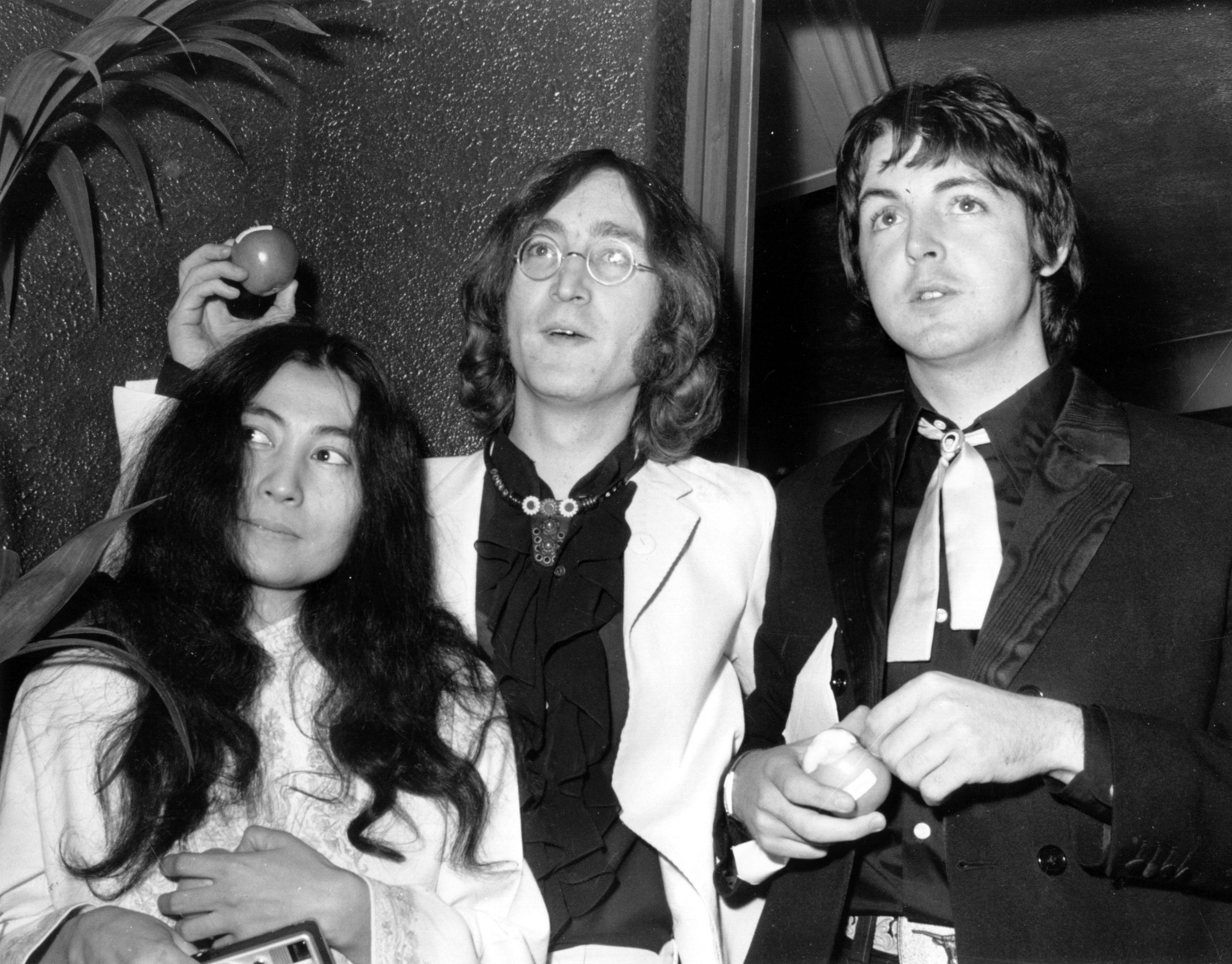 A black and white picture of Yoko Ono, John Lennon, and Paul McCartney. Lennon and McCartney hold apples.
