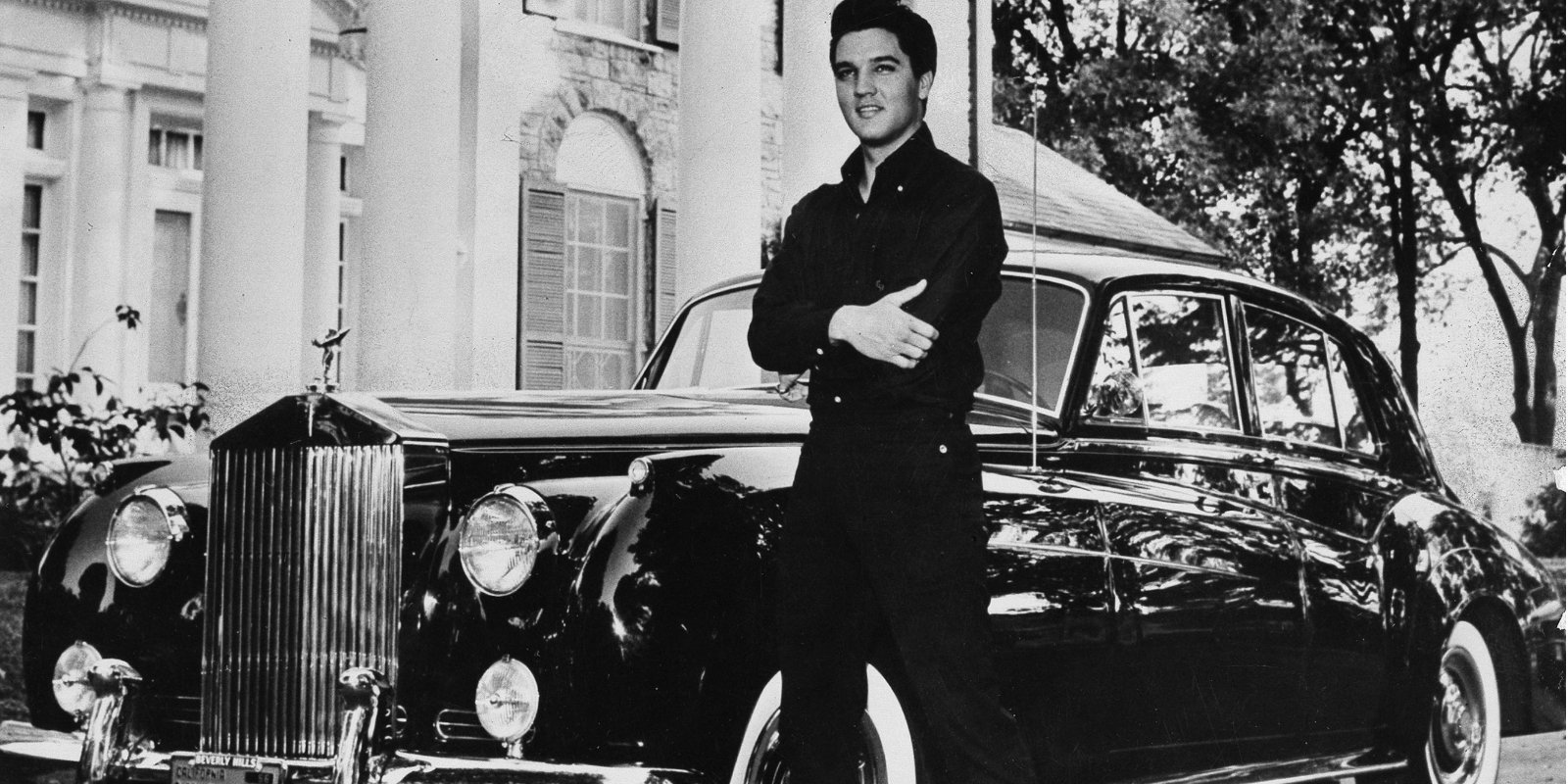 Elvis Presley poses in front of Graceland in a black and white image.