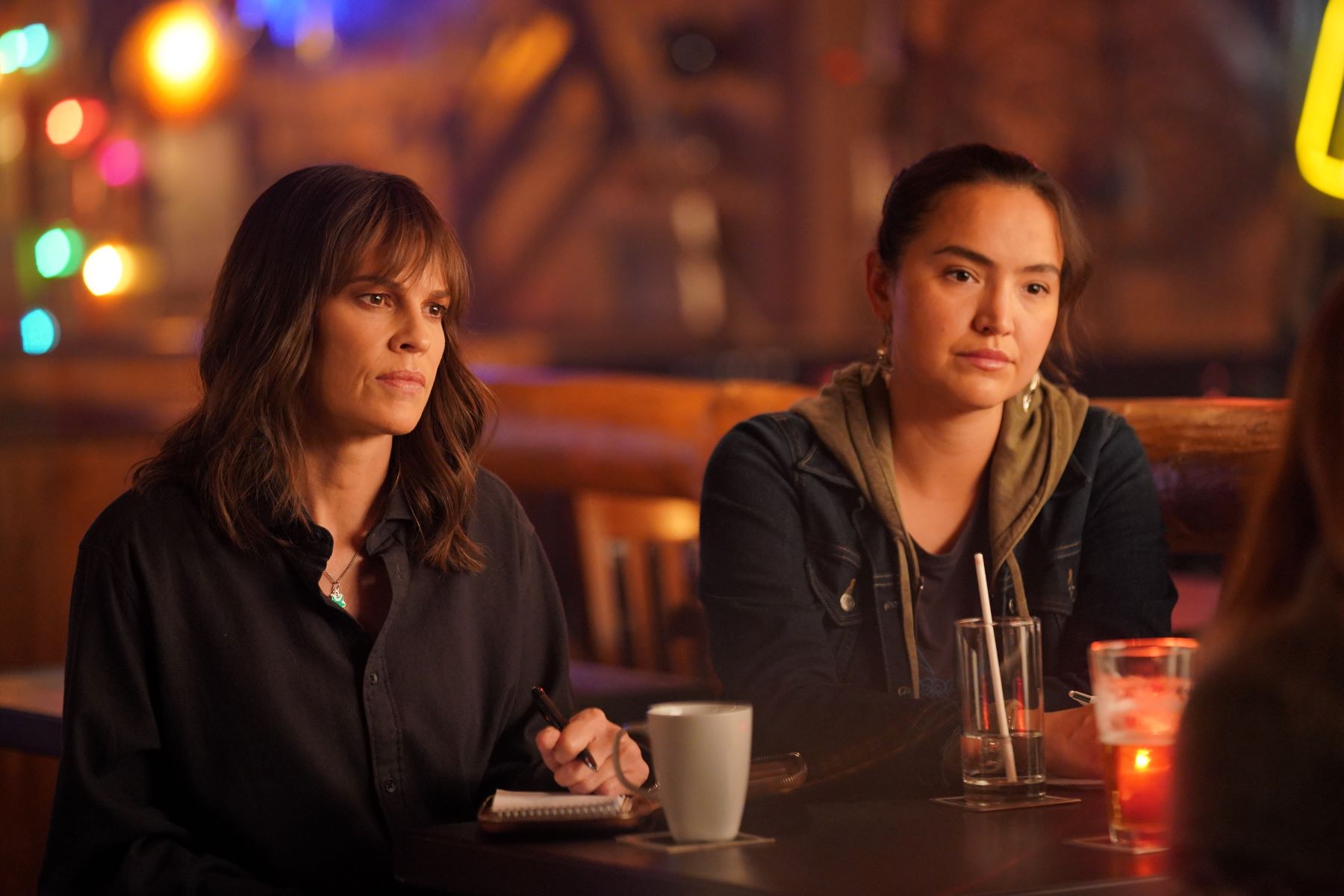 Hilary Swank and Grace Dove, in character as Eileen and Roz in 'Alaska Daily' Season 1 Episode 5, 'I Have No Idea What You’re Talking About, Eileen,' share a scene. Eileen wears a black button-up long-sleeved shirt. Roz wears a dark jean jacket over a brown hoodie and gray shirt.