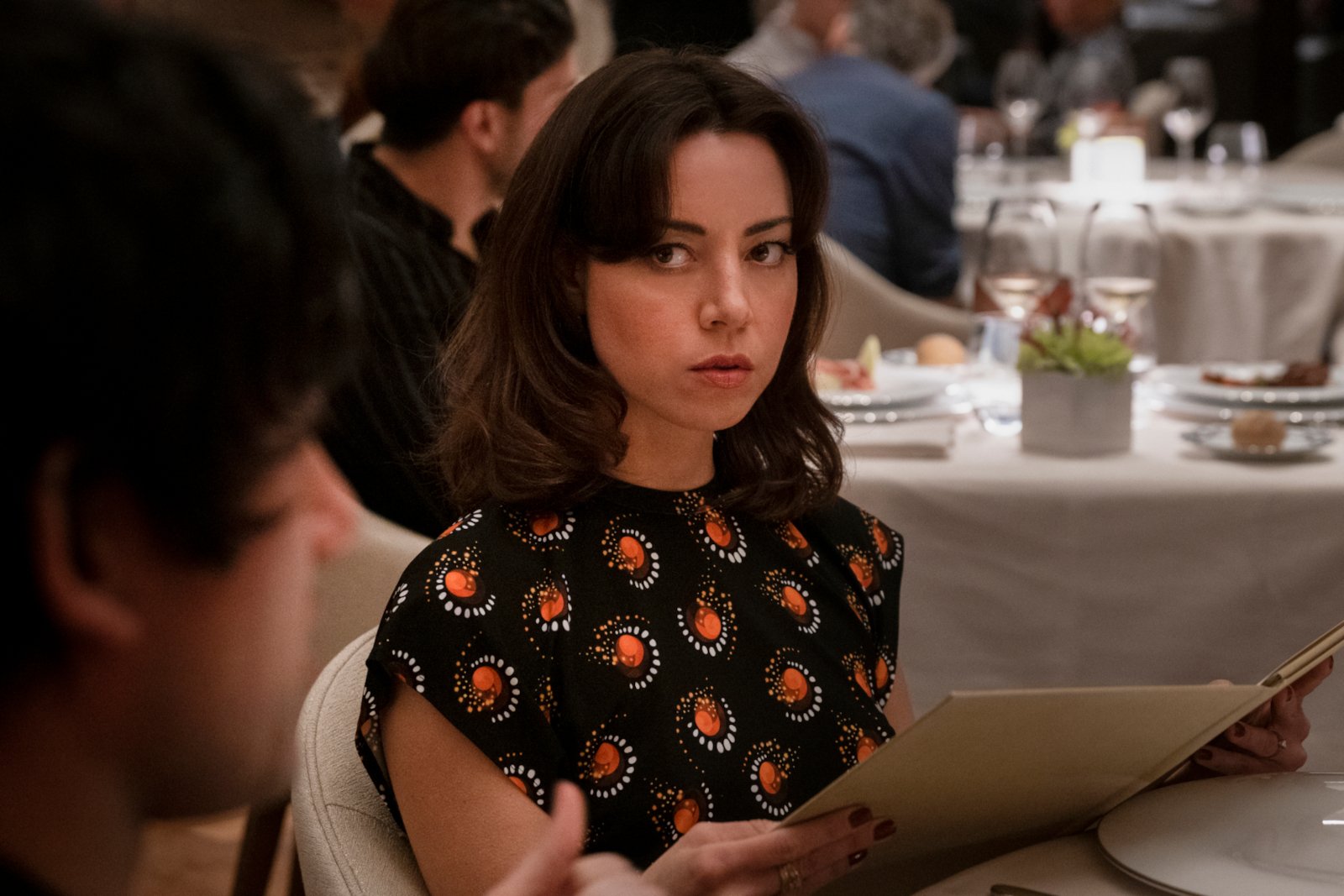Actor Aubrey Plaza as Harper Spiller in 'The White Lotus' Season 2. She's sitting at a table, holding a menu, and glaring at the person across from her.
